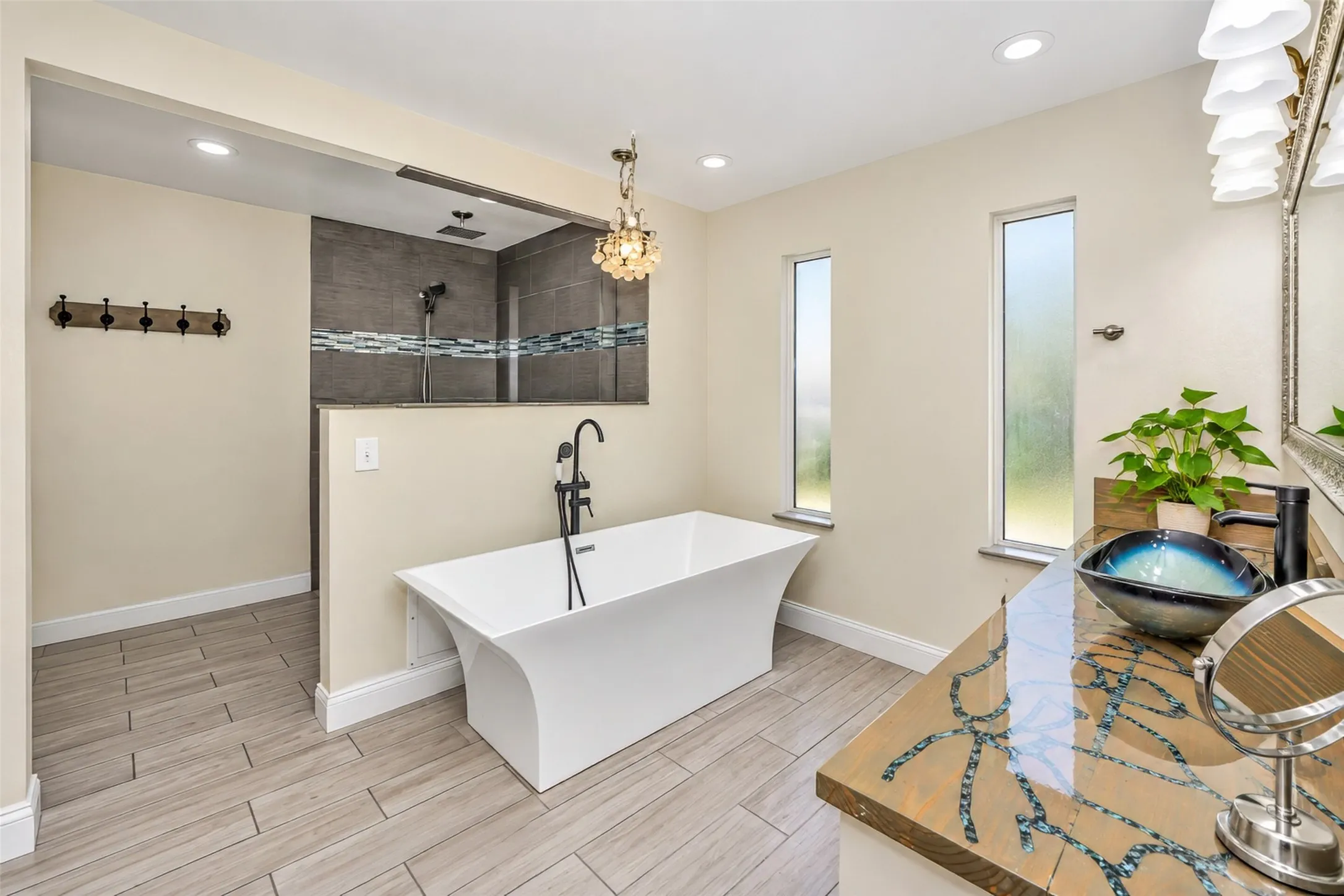 Freestanding soaking tub with a black floor-mounted faucet, walk-in shower with tile surround, wood-finish flooring, and a vanity with a vessel sink