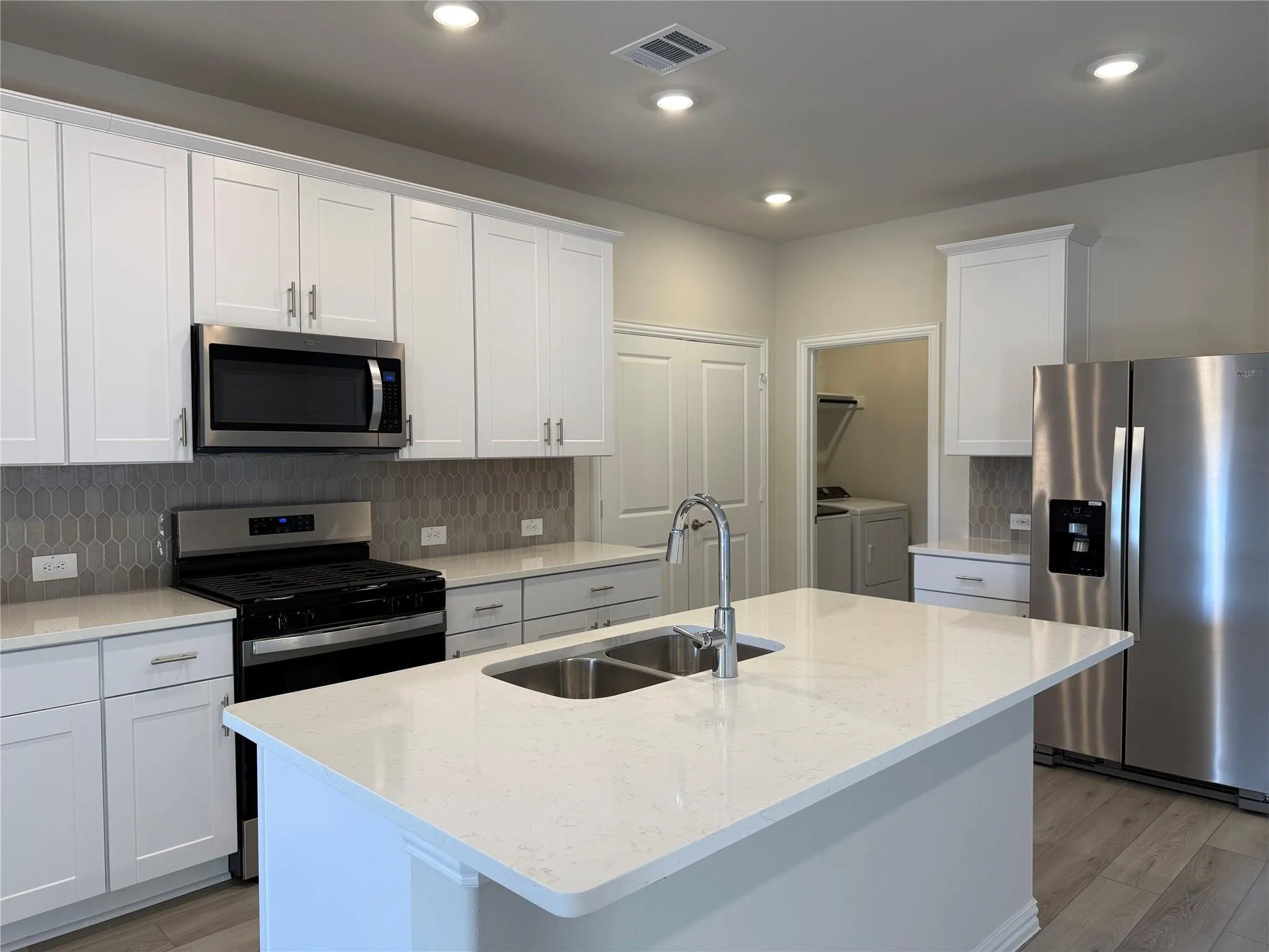 Kitchen featuring an island with a dual-basin sink, white shaker cabinetry, stainless steel appliances, and a gray tile backsplash