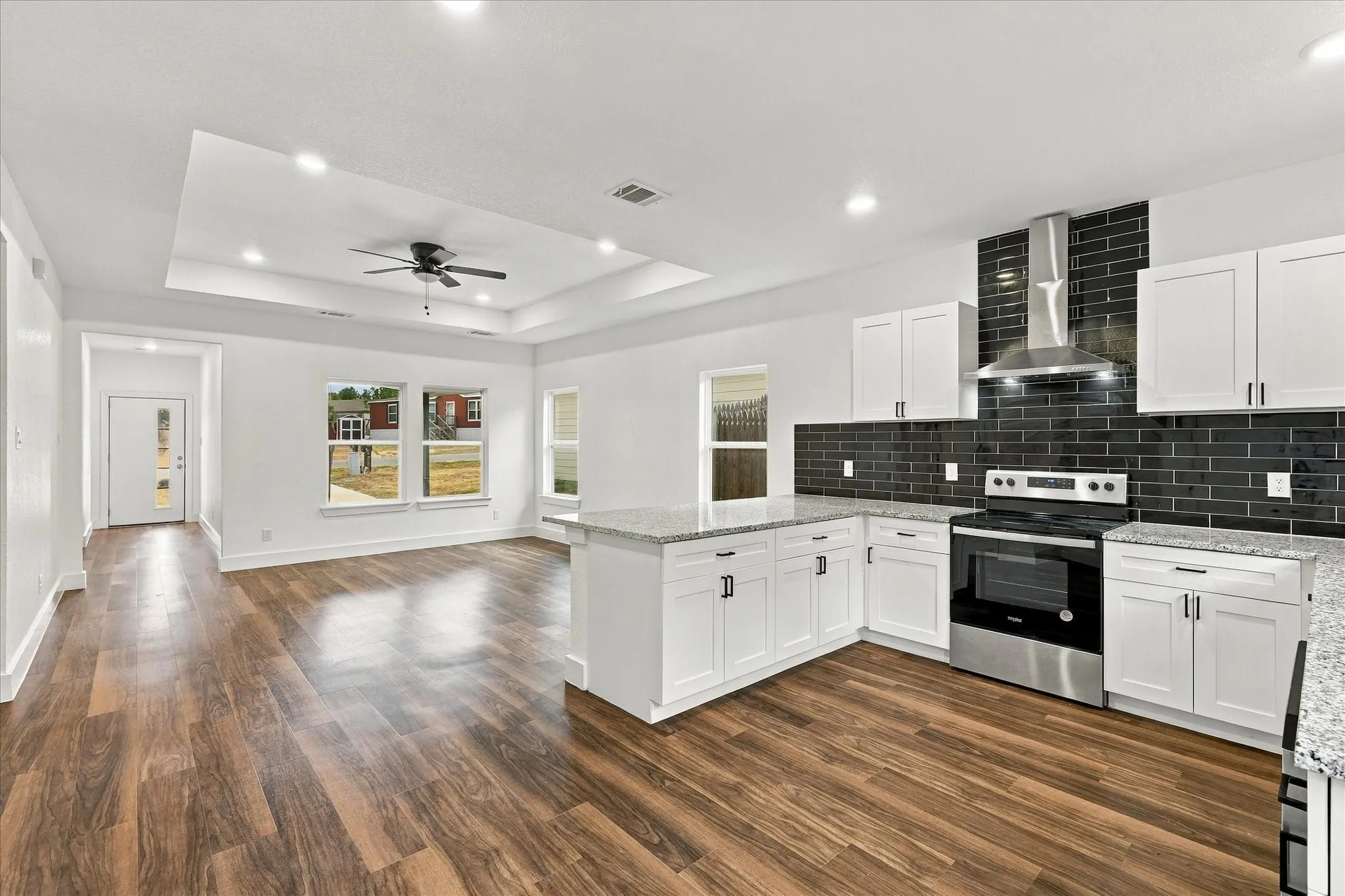 Open-concept living space featuring wood-finish flooring, a recessed ceiling with a ceiling fan, a kitchen island with stone countertops, white cabinetry with dark hardware, and a black subway tile backsplash