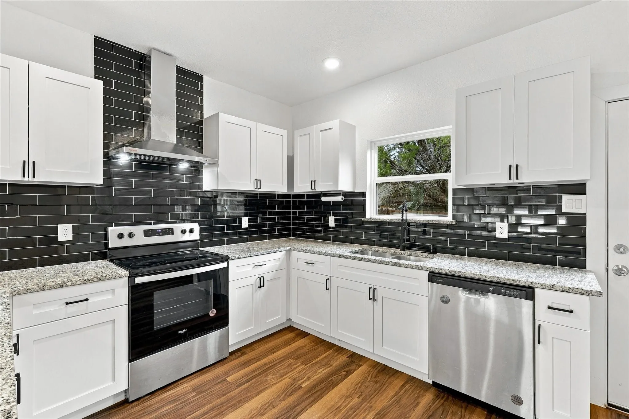 White shaker-style cabinetry, granite countertops, full black subway tile backsplash, stainless steel appliances, and wood-finish flooring