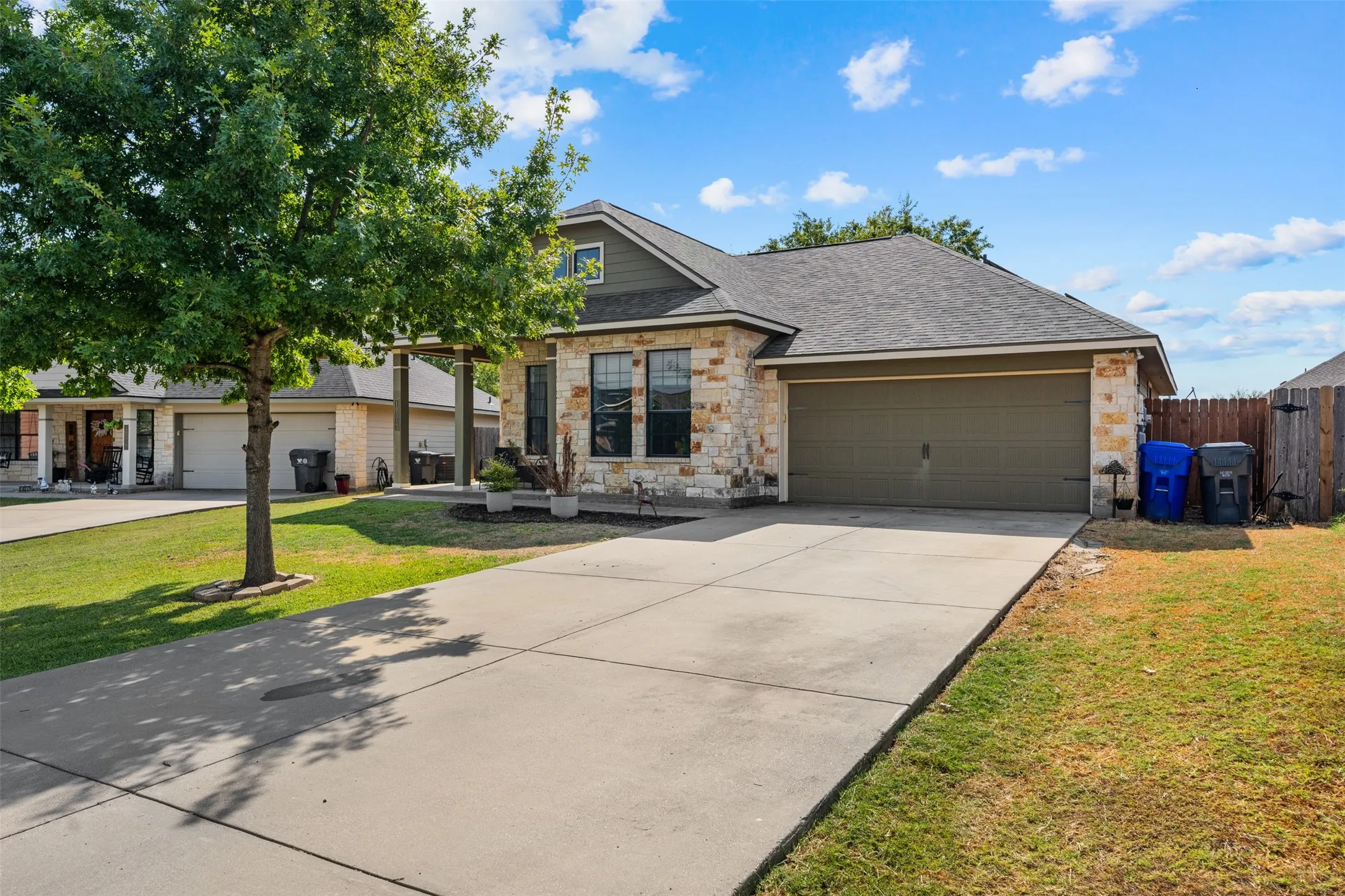 Stone veneer exterior with dark shingle roof, attached garage with a solid door, concrete driveway, and a front yard with mature trees