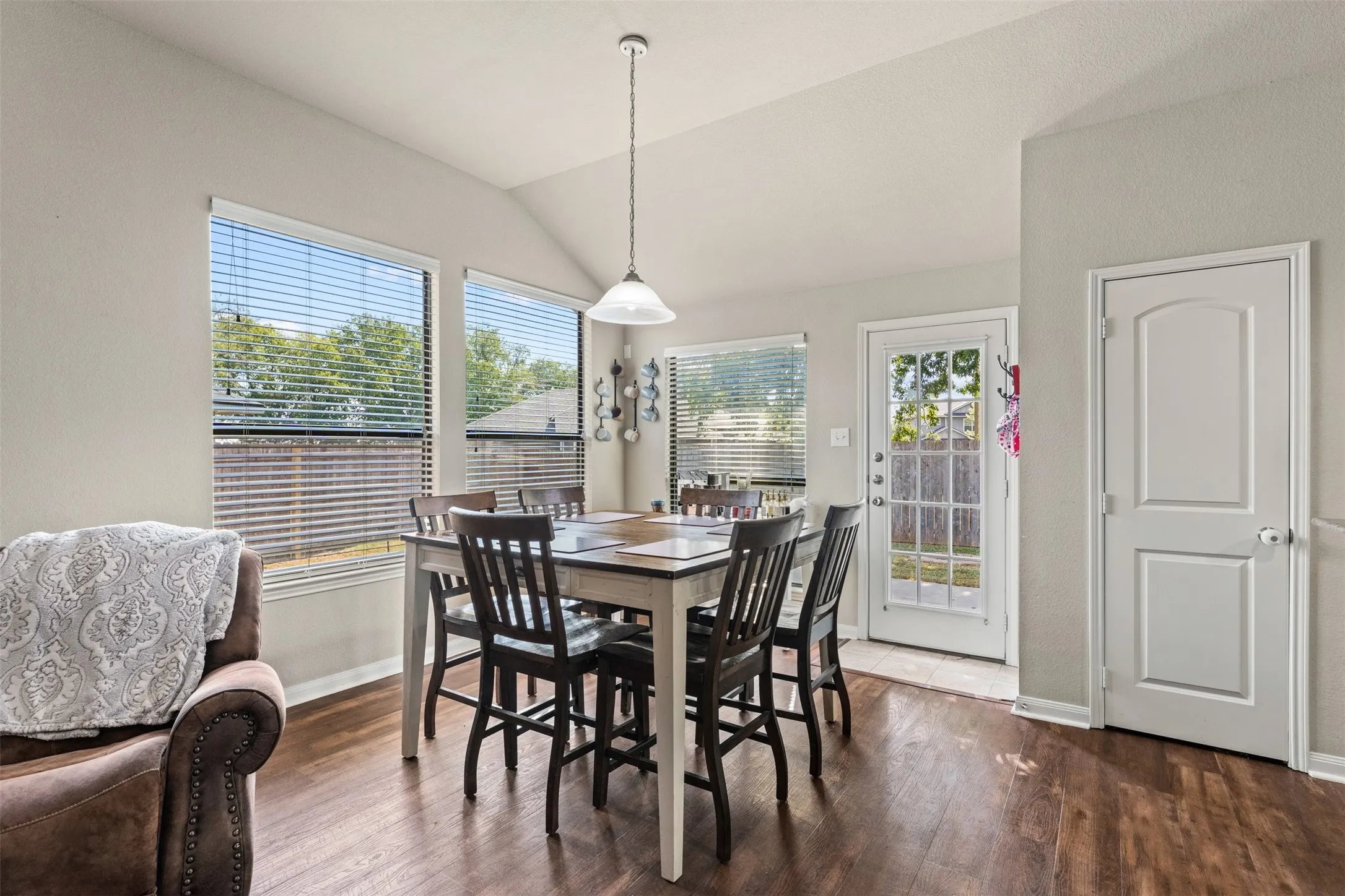 Vaulted ceiling with a pendant light fixture, wood-finish flooring, multiple windows with blinds, and a paneled door