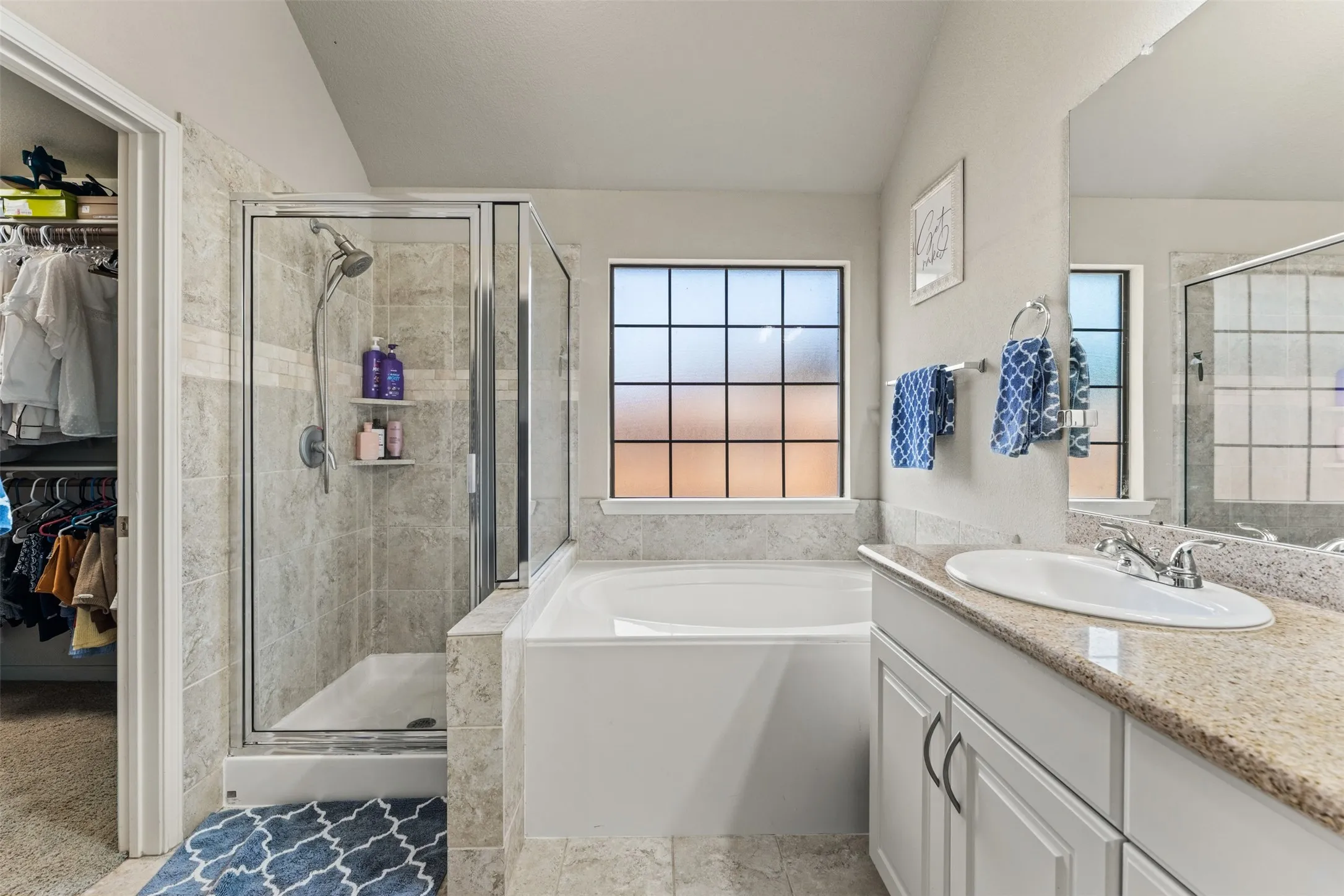 Built-in soaking tub, glass-enclosed shower with tile surround, single vanity with countertop, and framed window