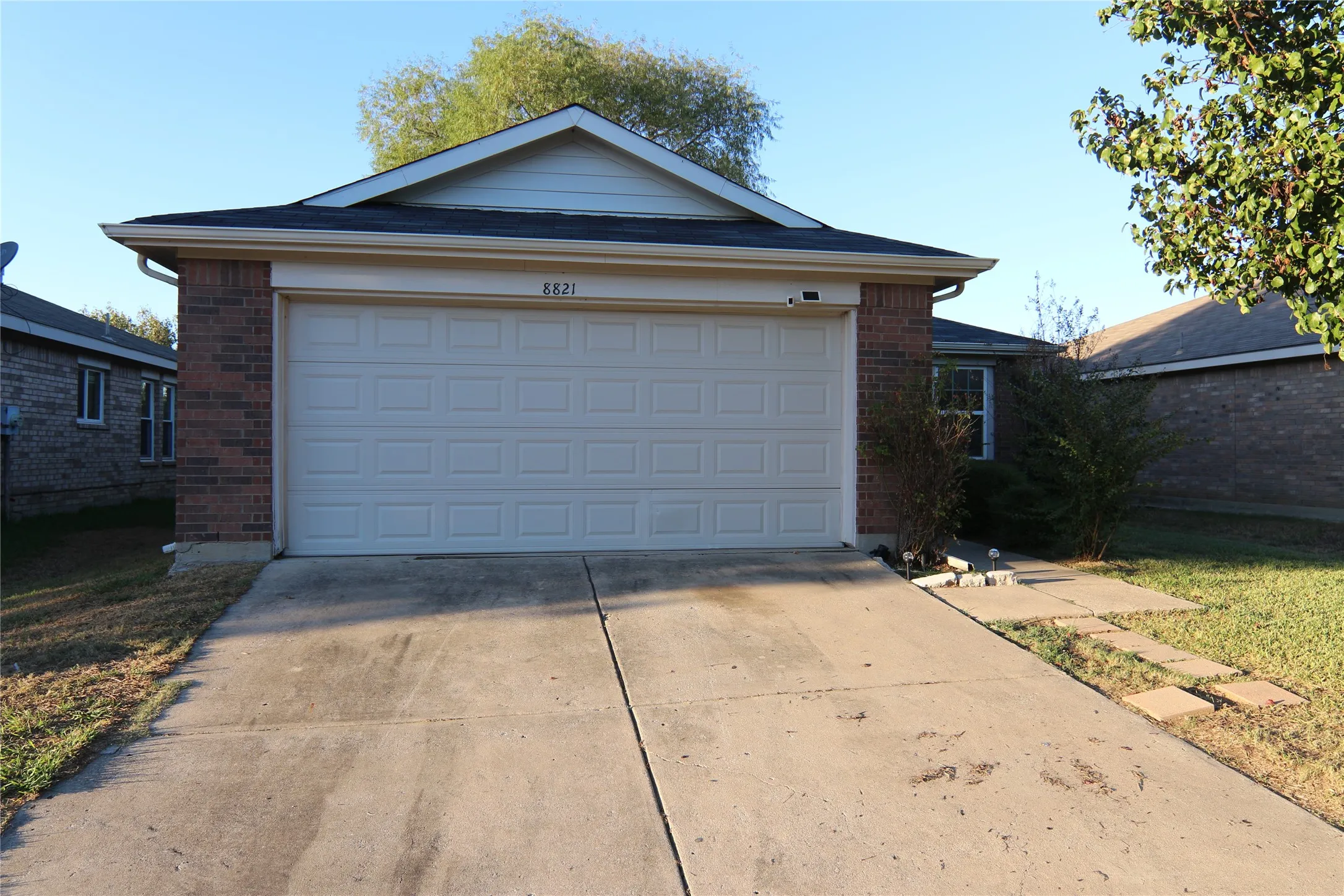Brick exterior with an attached garage, concrete driveway, and established landscaping