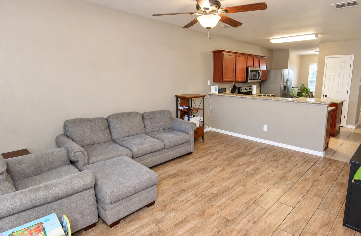 Open-concept living space featuring wood-finish flooring, a ceiling fan with integrated lighting, a kitchen pass-through with a countertop, and wood cabinetry
