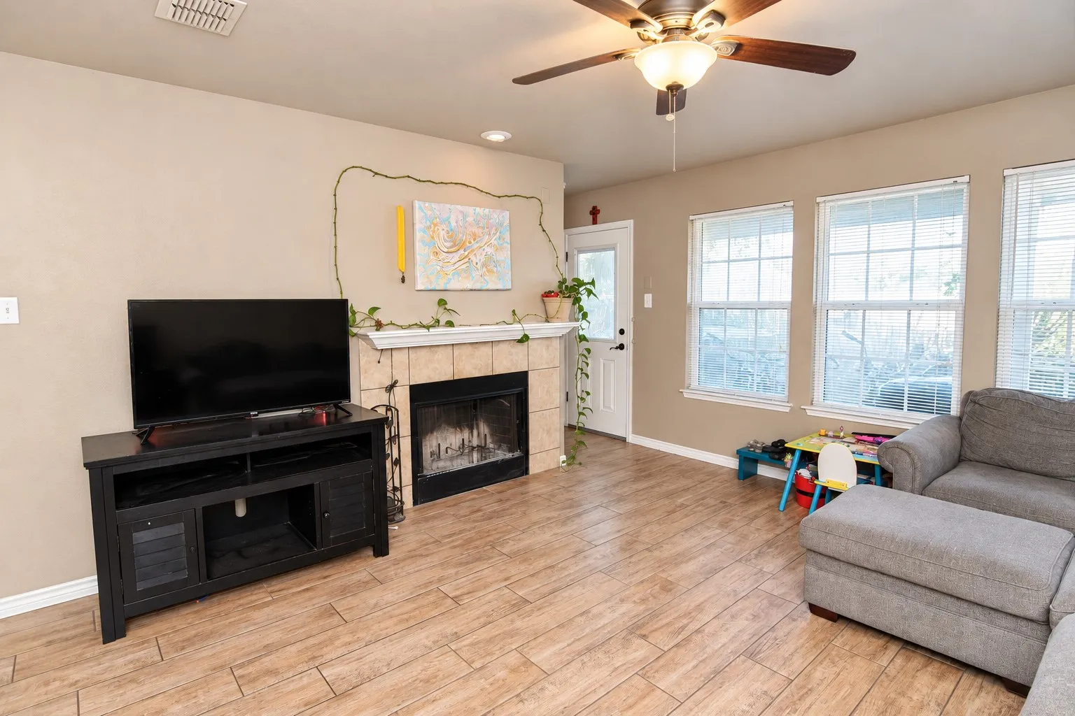 Living area featuring wood-finish flooring, a tiled fireplace with a mantel, multiple windows, and a ceiling fan with integrated lighting