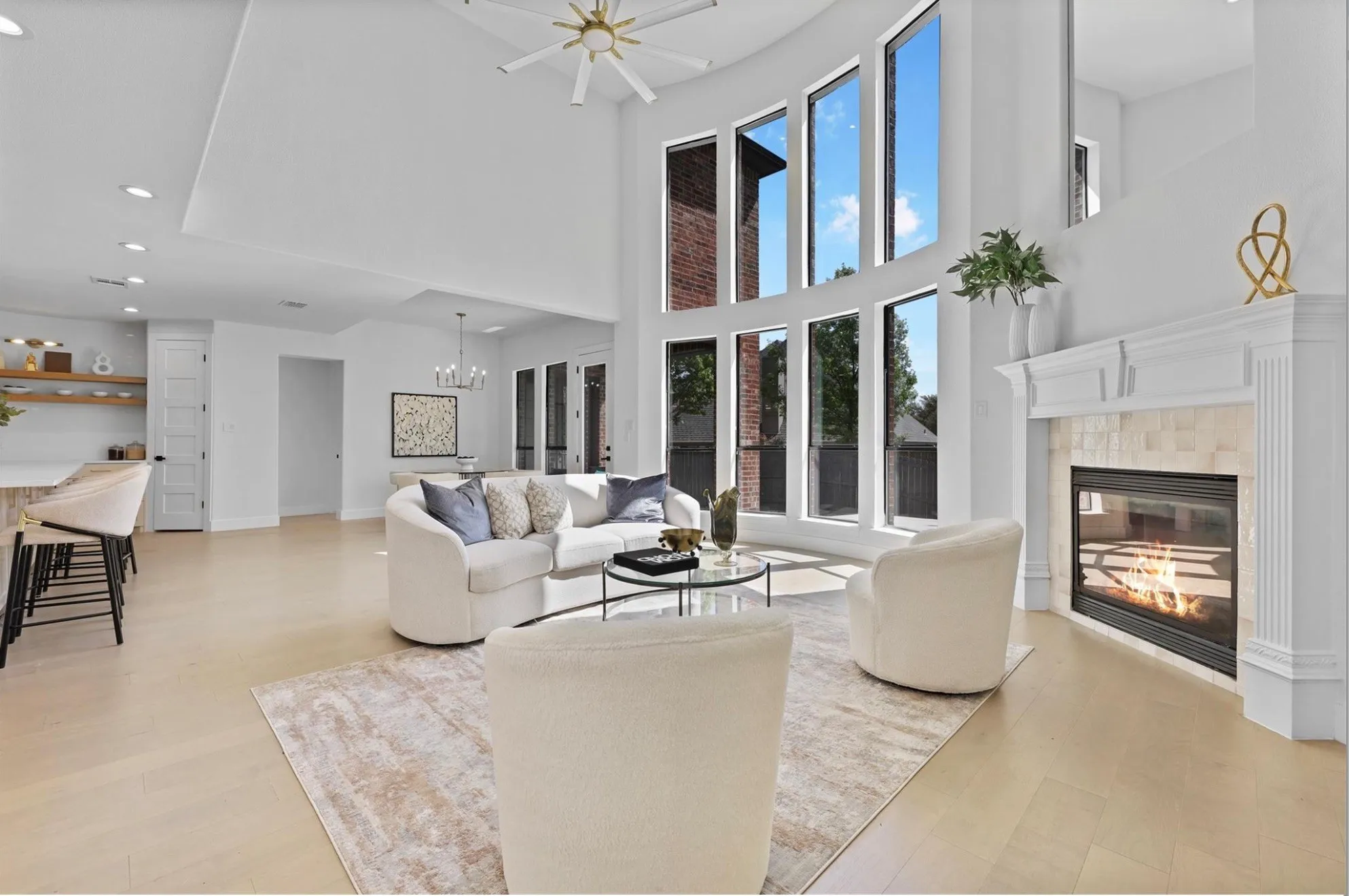 Double-height ceiling, floor-to-ceiling windows, wood-finish flooring, built-in fireplace with stone surround, and a ceiling fan with integrated lighting