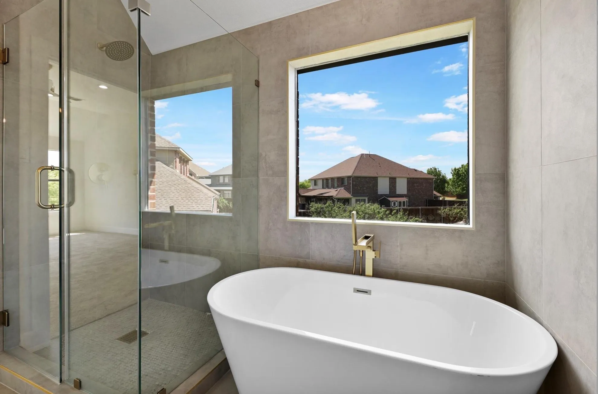 Freestanding soaking tub with a gold-tone faucet, walk-in glass shower, and large window