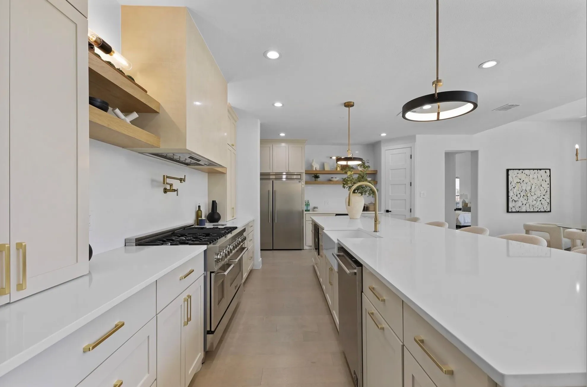 Expansive kitchen featuring a large central island with a sink, stainless steel appliances, white cabinetry with gold-finish hardware, and recessed lighting