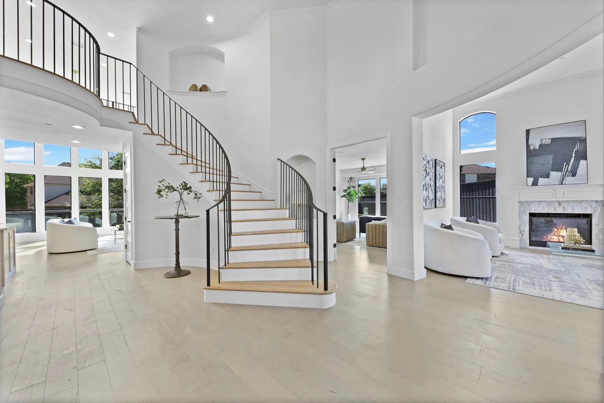 Grand curved staircase with wrought iron balusters, high ceilings, large arched windows, wood-finish flooring, and a fireplace with a decorative mantel
