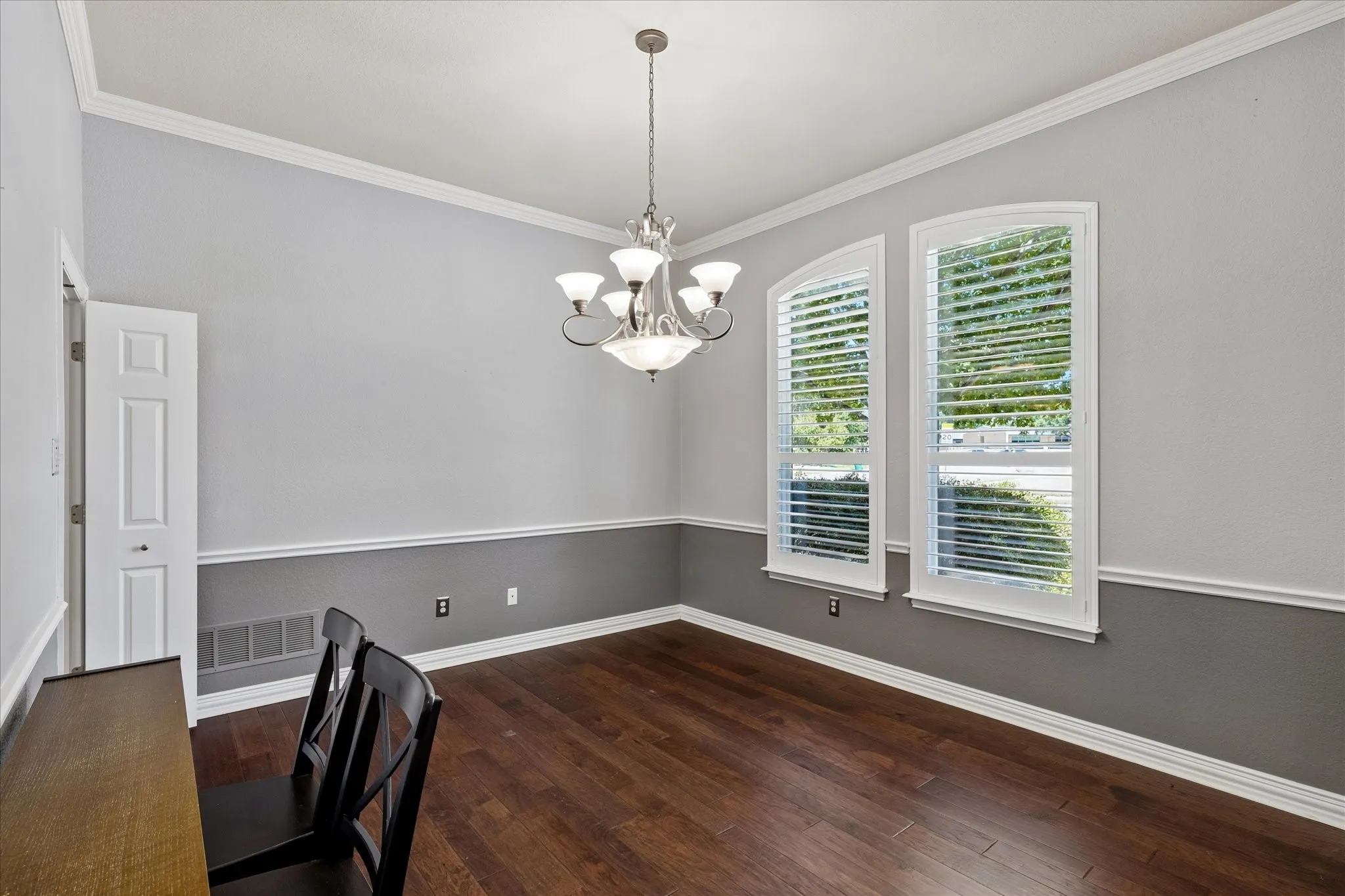 Wood-finish flooring, two-tone wall treatment with chair rail, decorative crown molding, two arched windows with plantation shutters, and a five-light chandelier