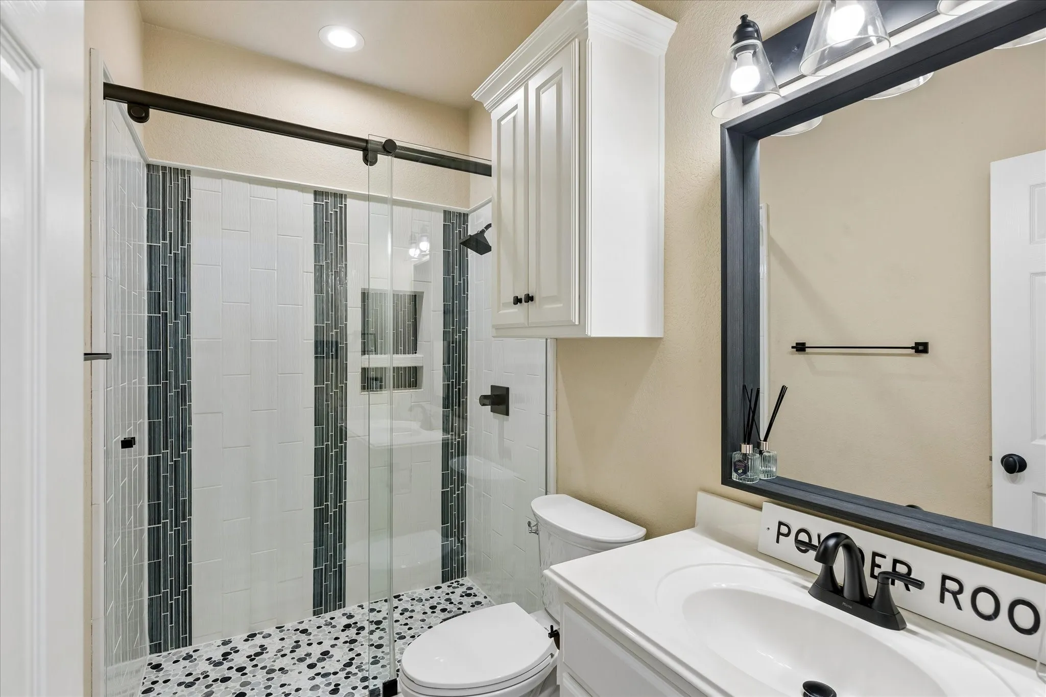 Glass-enclosed shower with subway tile and mosaic accents, pebble-style shower floor, white vanity with integrated sink, framed vanity mirror, and overhead cabinet storage
