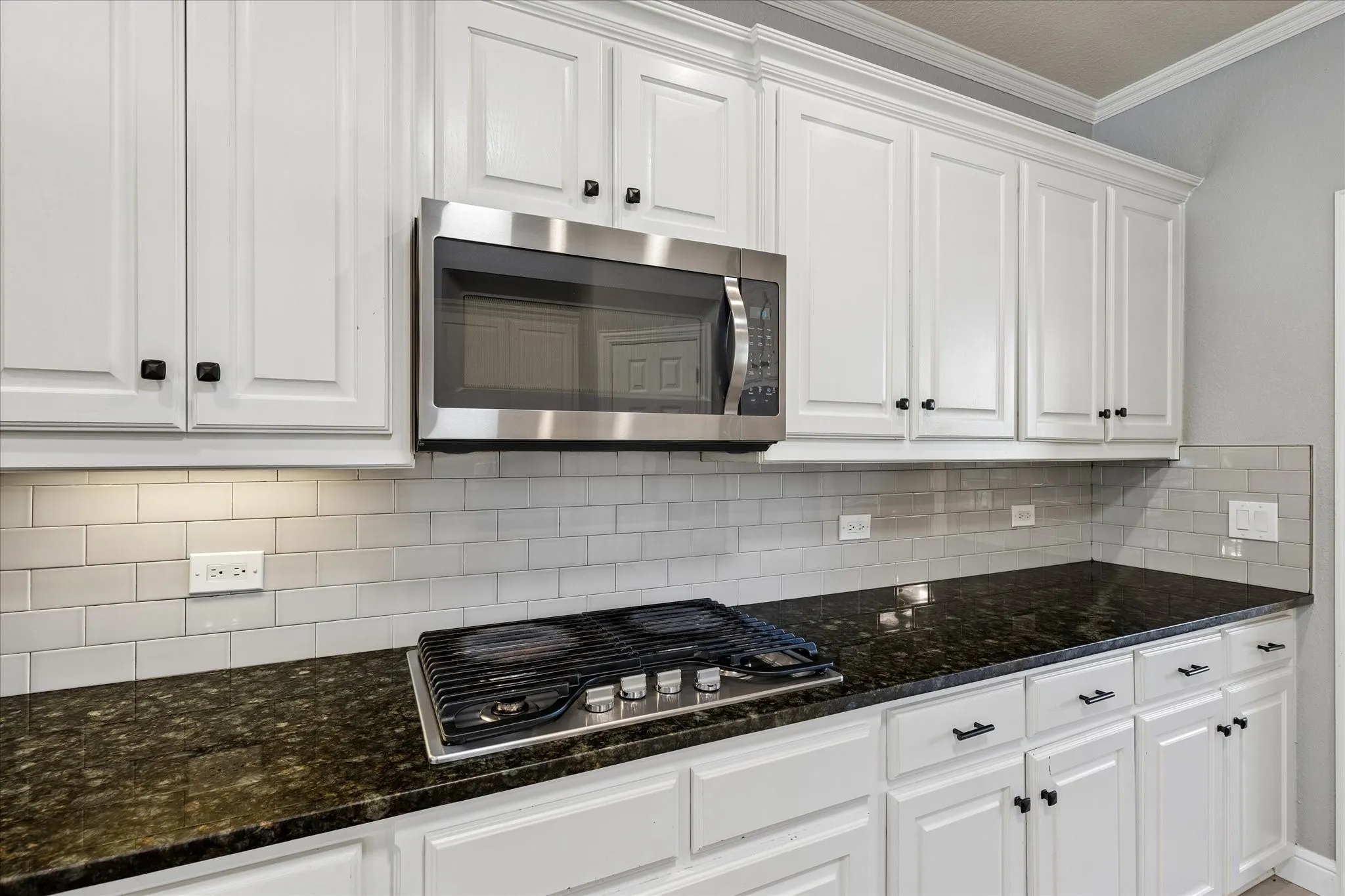 White cabinetry, dark stone countertops, stainless steel microwave, gas cooktop, and subway tile backsplash