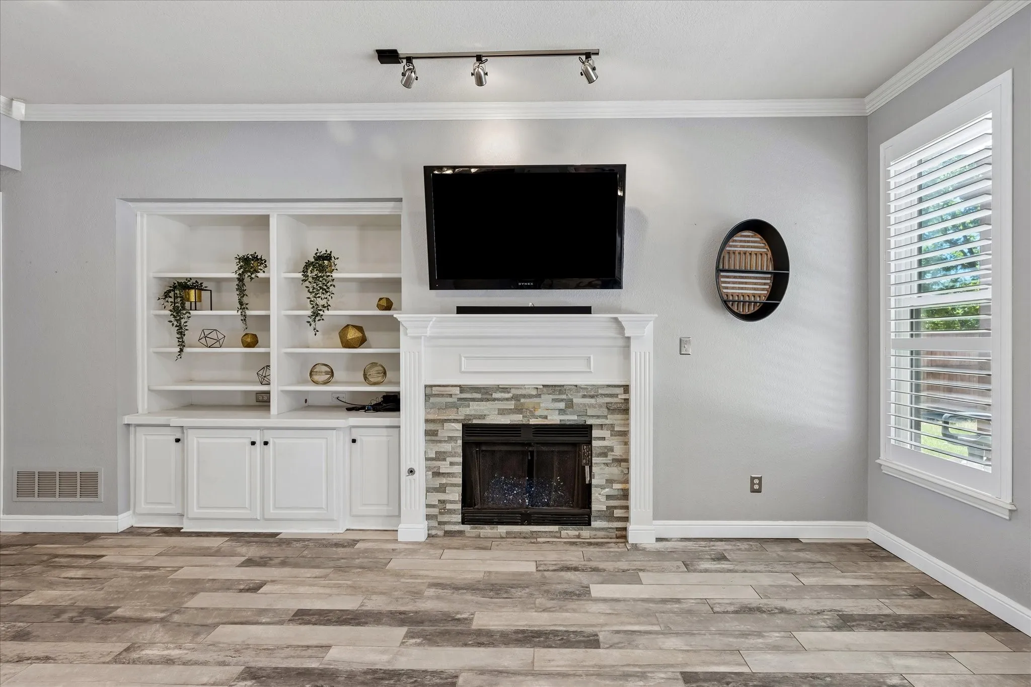 Built-in shelving and cabinetry, fireplace with stone surround, crown molding, wood-finish flooring, and window with plantation shutters