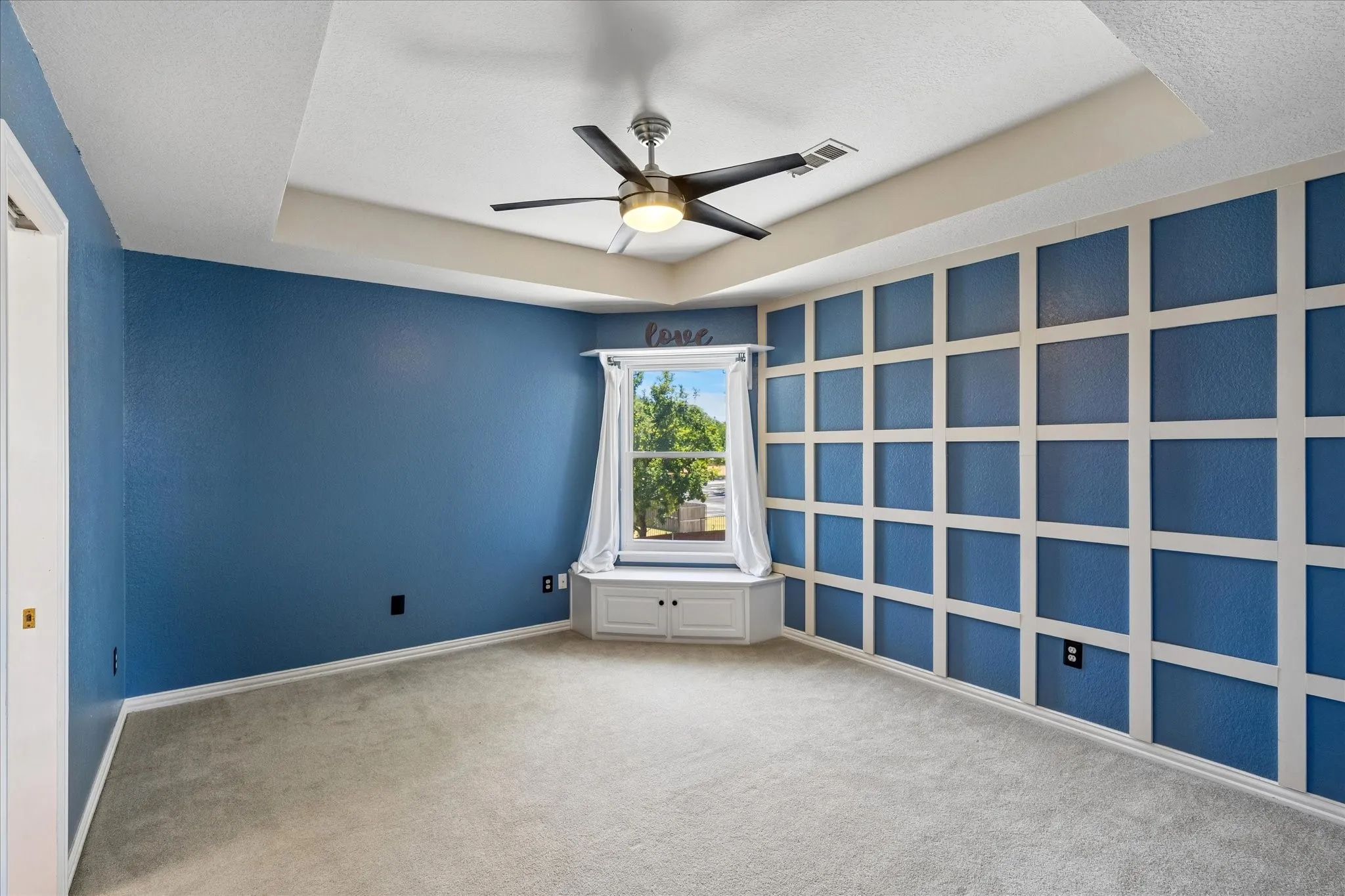 Tray ceiling with a ceiling fan, accent wall with decorative paneling, built-in window bench, and wall-to-wall carpeting