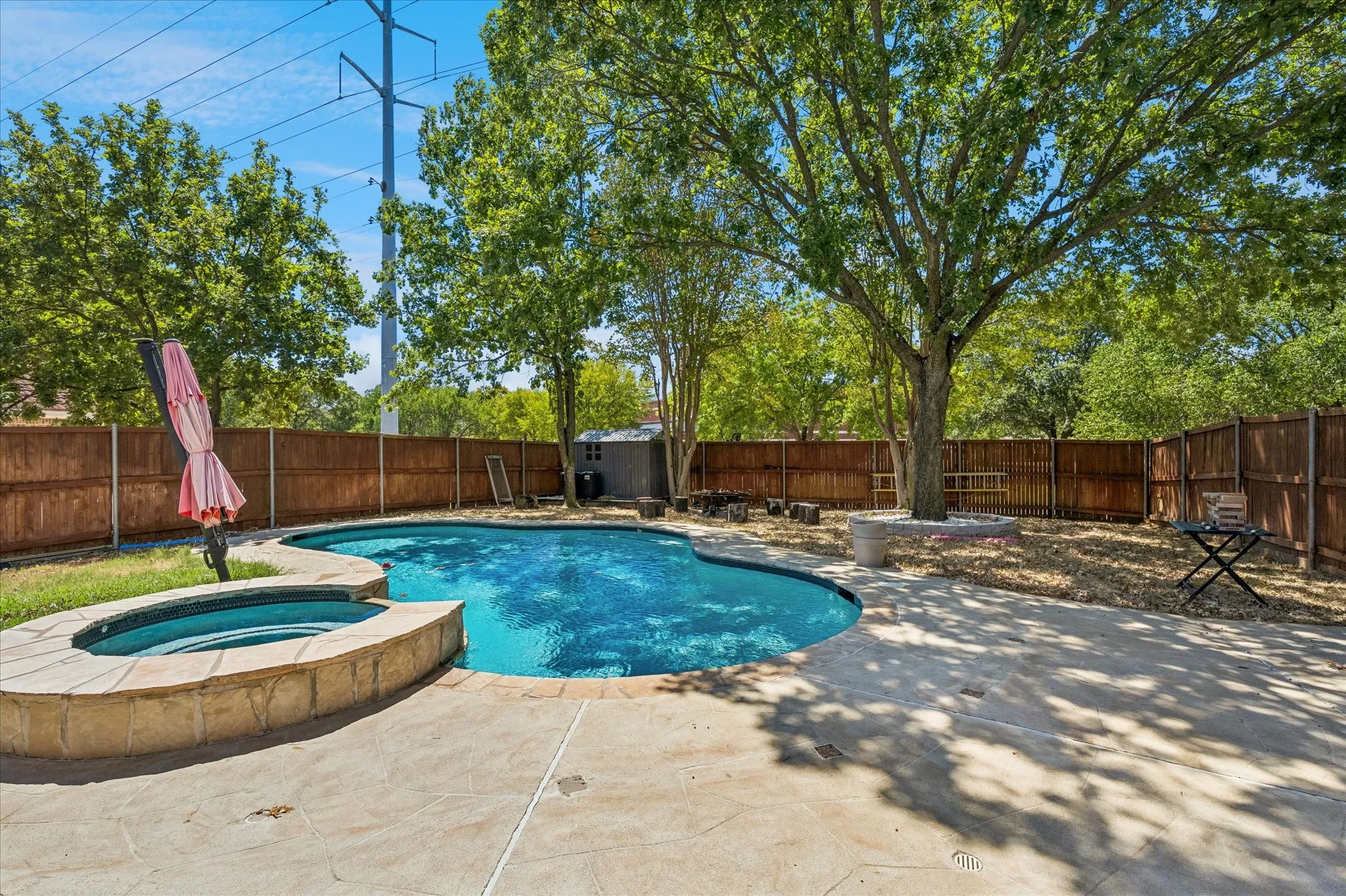 In-ground pool with integrated spa, concrete decking, mature trees, wood privacy fencing, and an outdoor storage shed