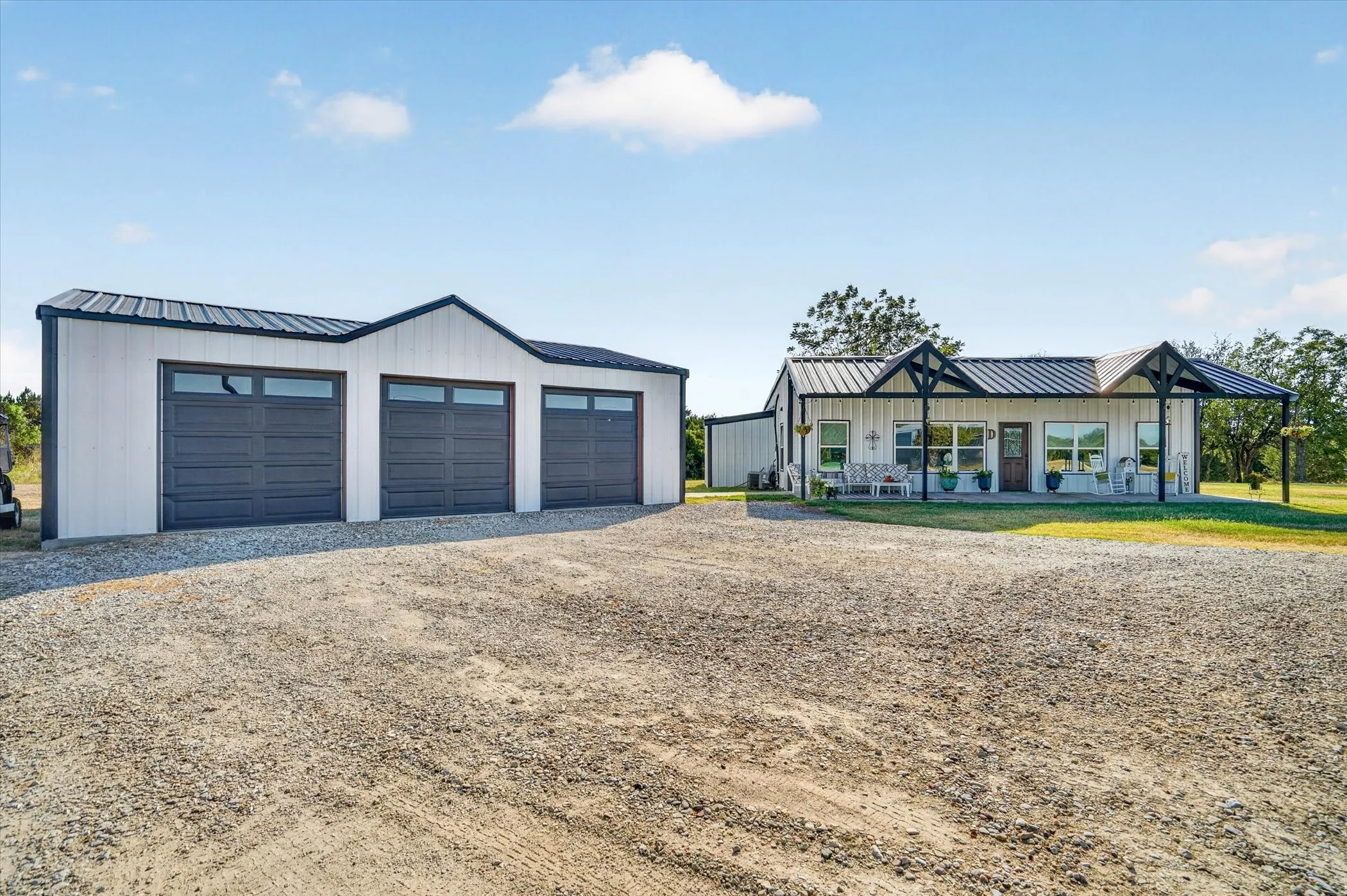 White exterior with black trim, covered front porch, gravel driveway, and a detached three-car garage