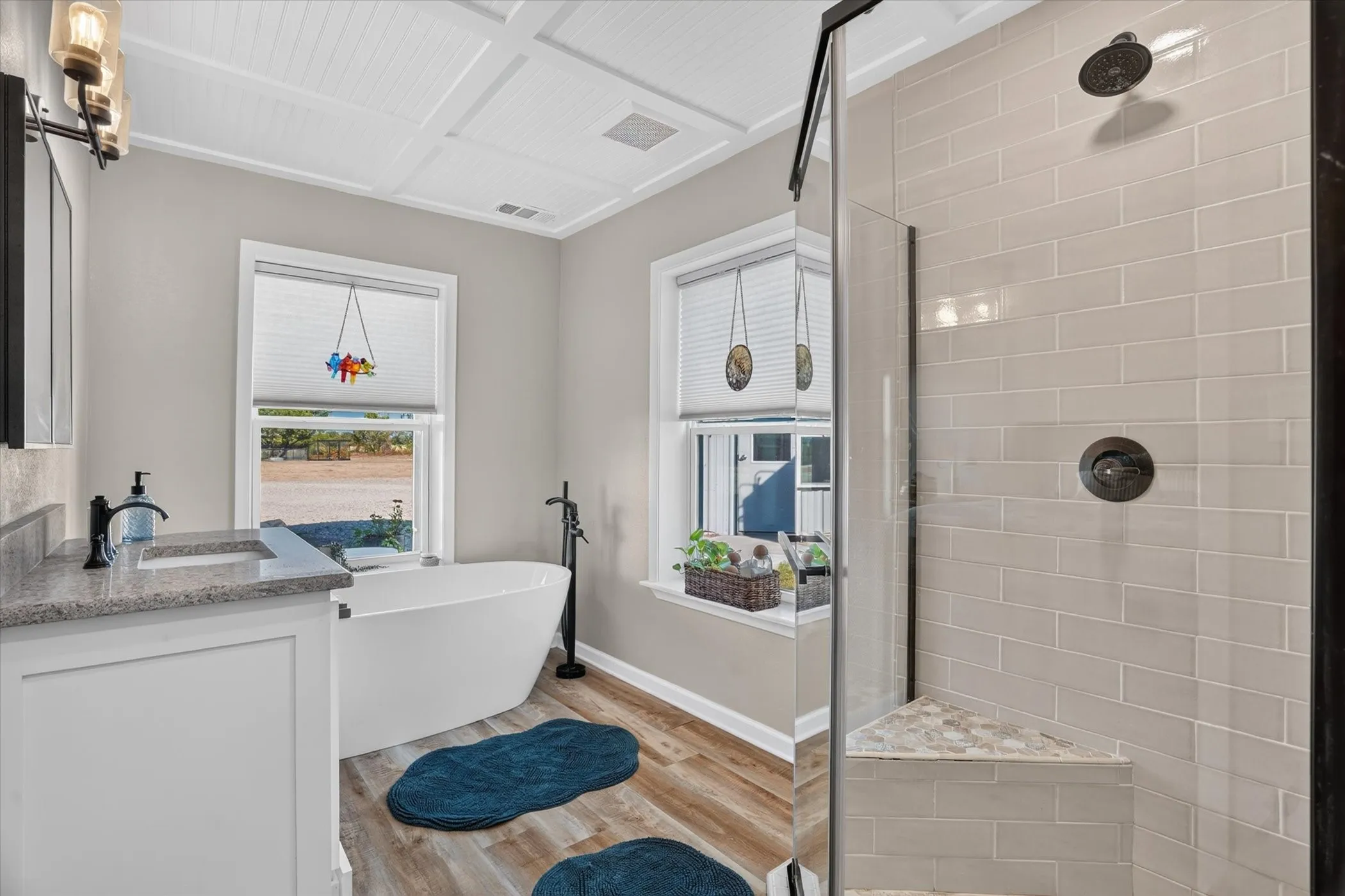 White coffered ceiling, freestanding soaking tub with floor-mounted faucet, wood-finish flooring, vanity with stone countertop, and a glass-enclosed shower with subway tile