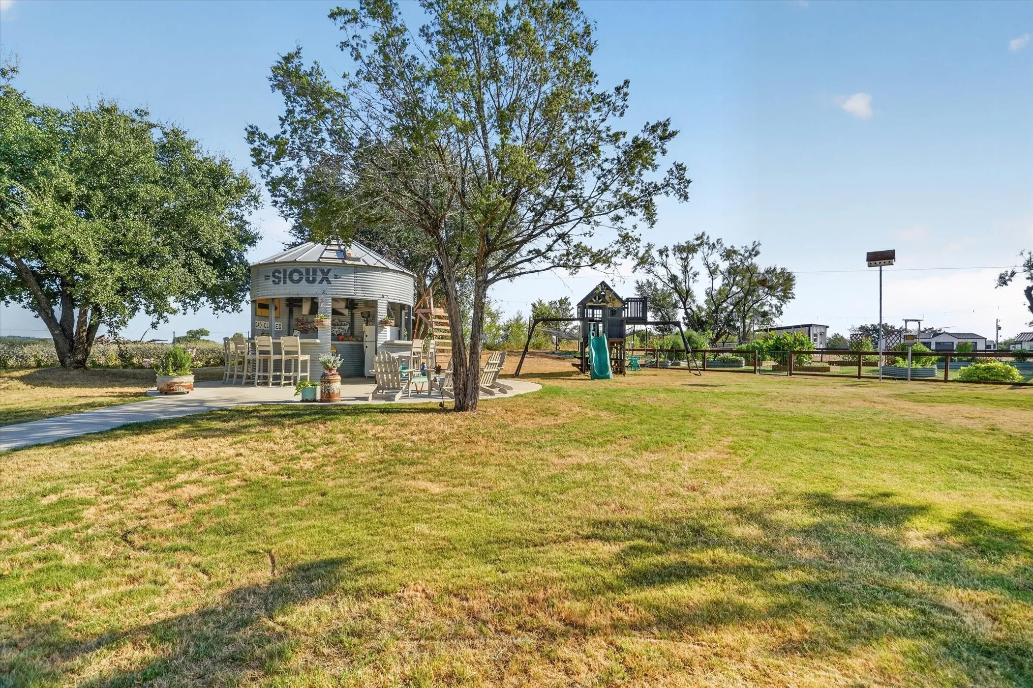 Expansive outdoor area featuring a circular concrete patio with a built-in bar structure, a separate play structure with a slide and swings, and mature trees providing shade