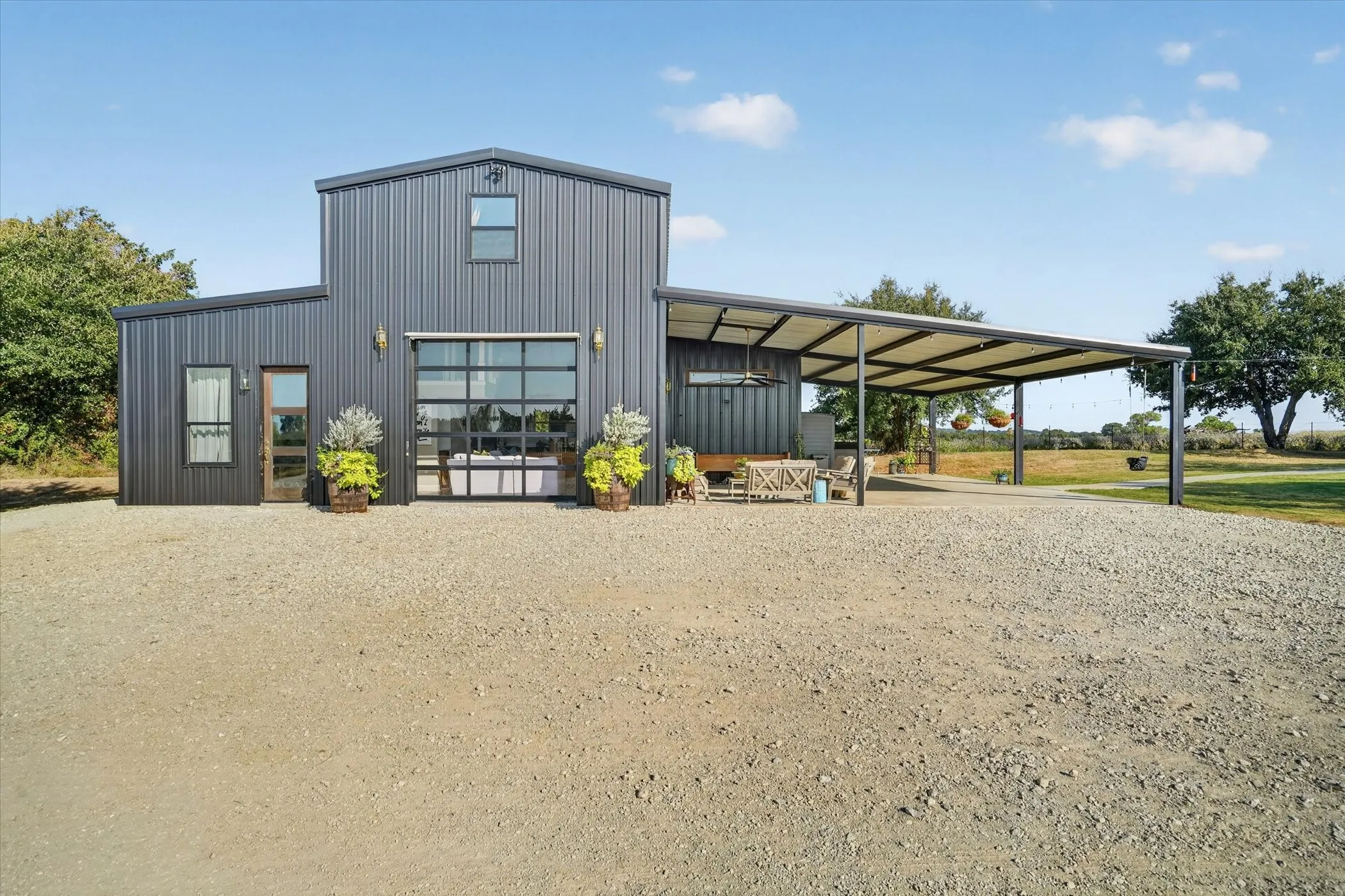 Modern metal exterior with a two-story structure, expansive covered patio, large glass panel garage door, and a gravel driveway