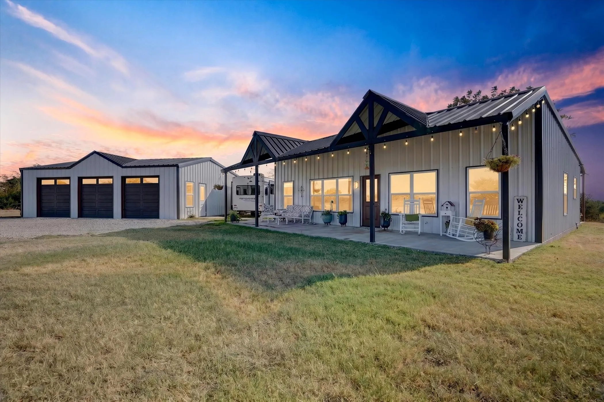 Metal-sided residence featuring a covered front porch with string lighting, multiple windows, and a separate garage structure with three bays