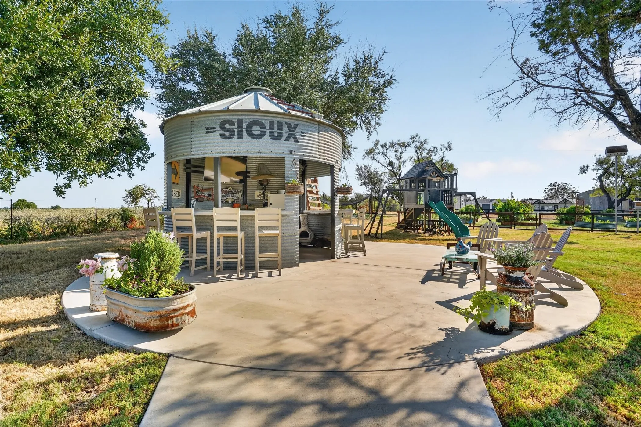 Expansive concrete patio featuring a unique grain silo bar, mature trees, and a wooden playground structure