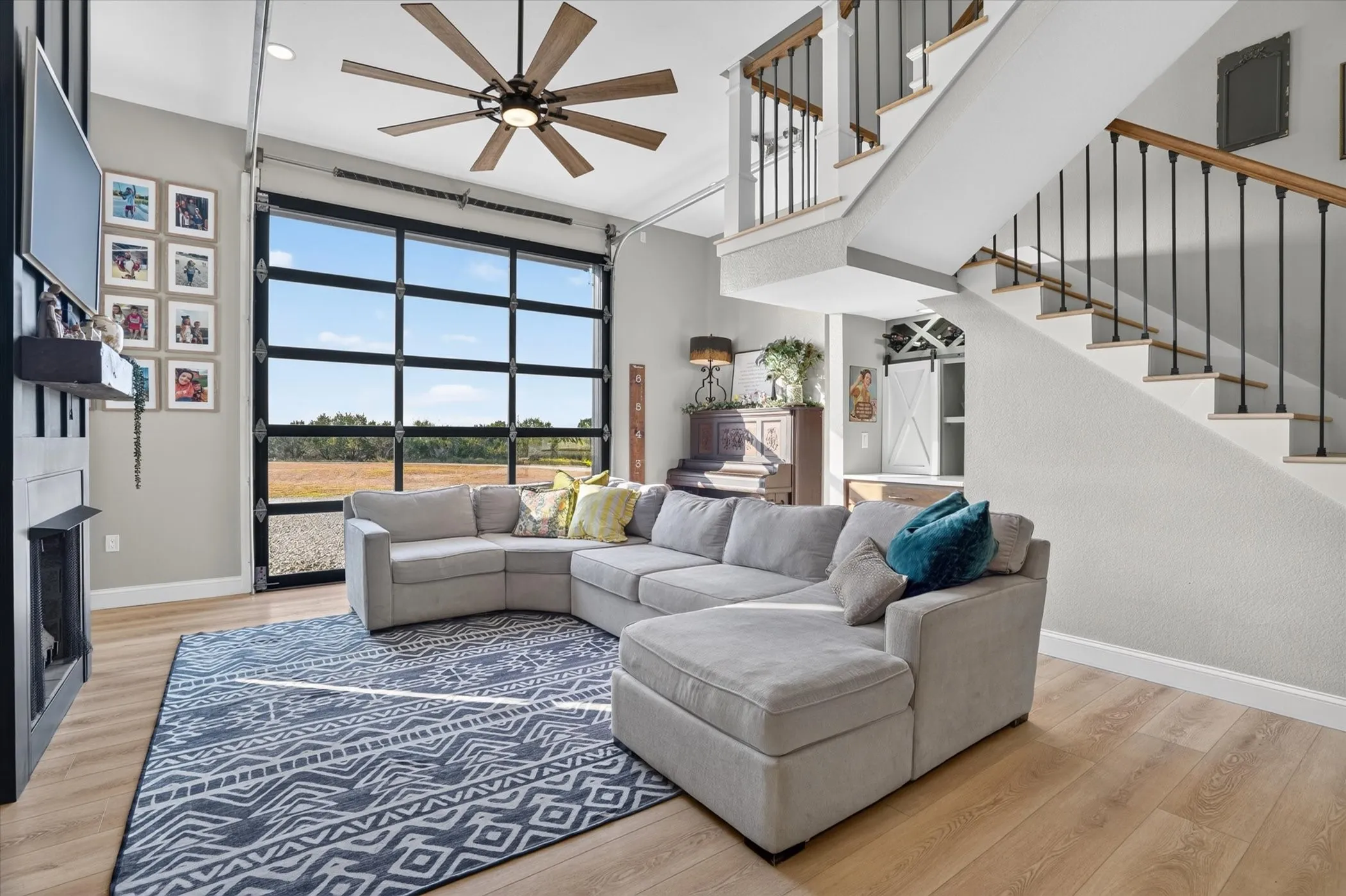 Expansive glass panel door, wood-finish flooring, two-story ceiling, black metal and wood staircase, and a prominent ceiling fan