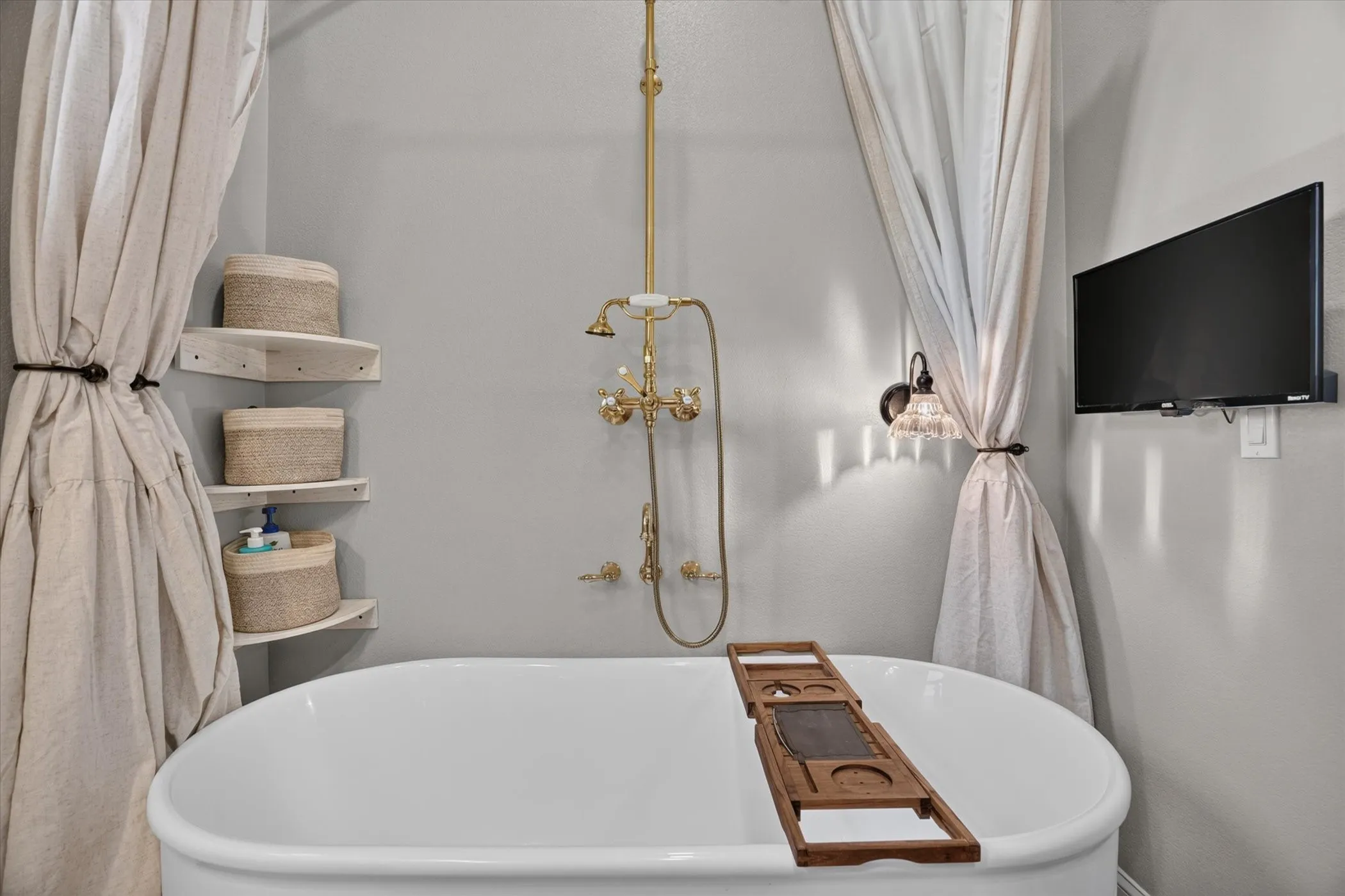 Freestanding bathtub with a floor-mounted faucet and showerhead, corner shelving, and a wall-mounted light fixture
