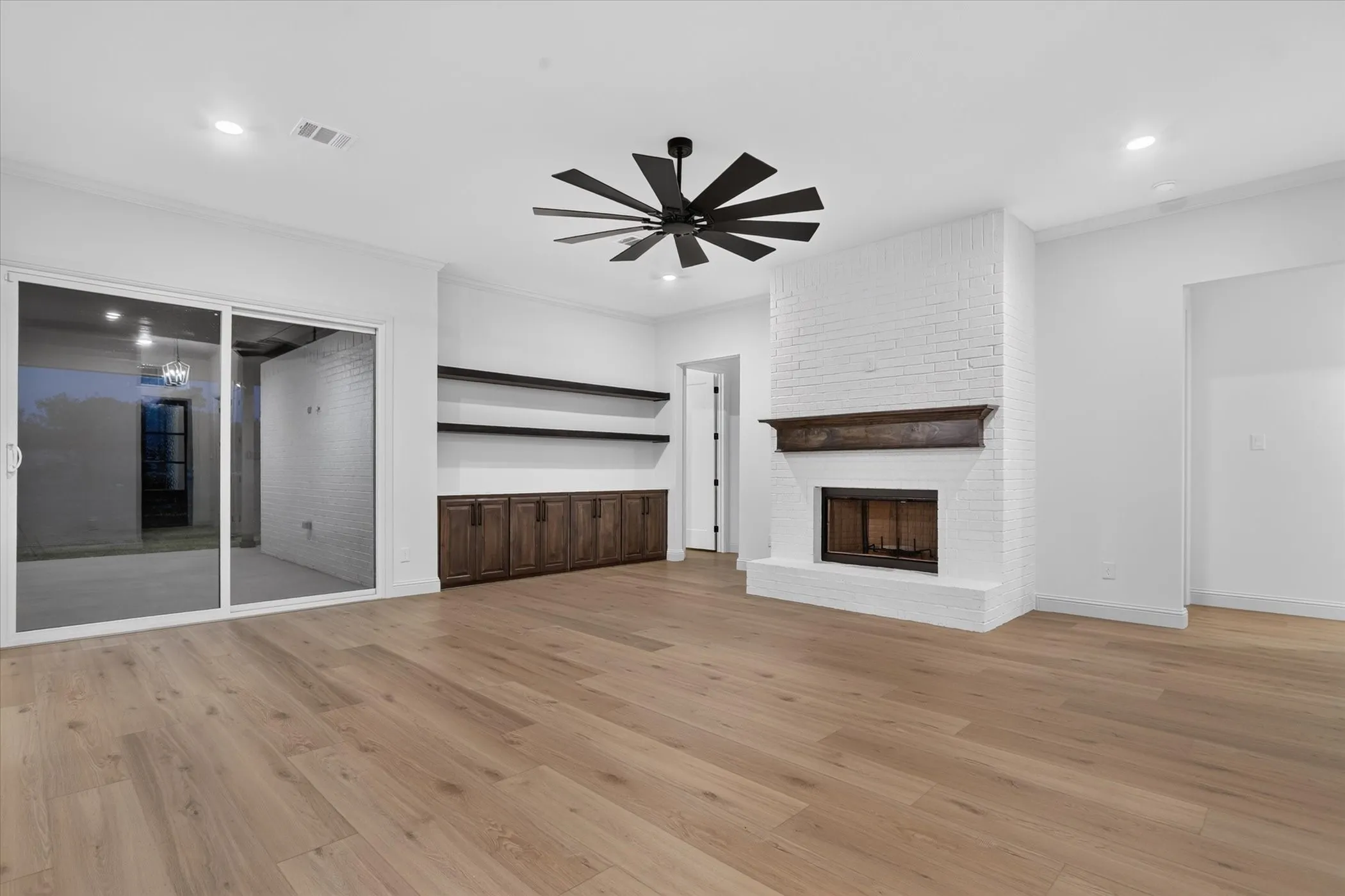 Wood-finish flooring, white brick fireplace with dark wood mantel, built-in cabinetry with open shelving, sliding glass door, and ceiling fan