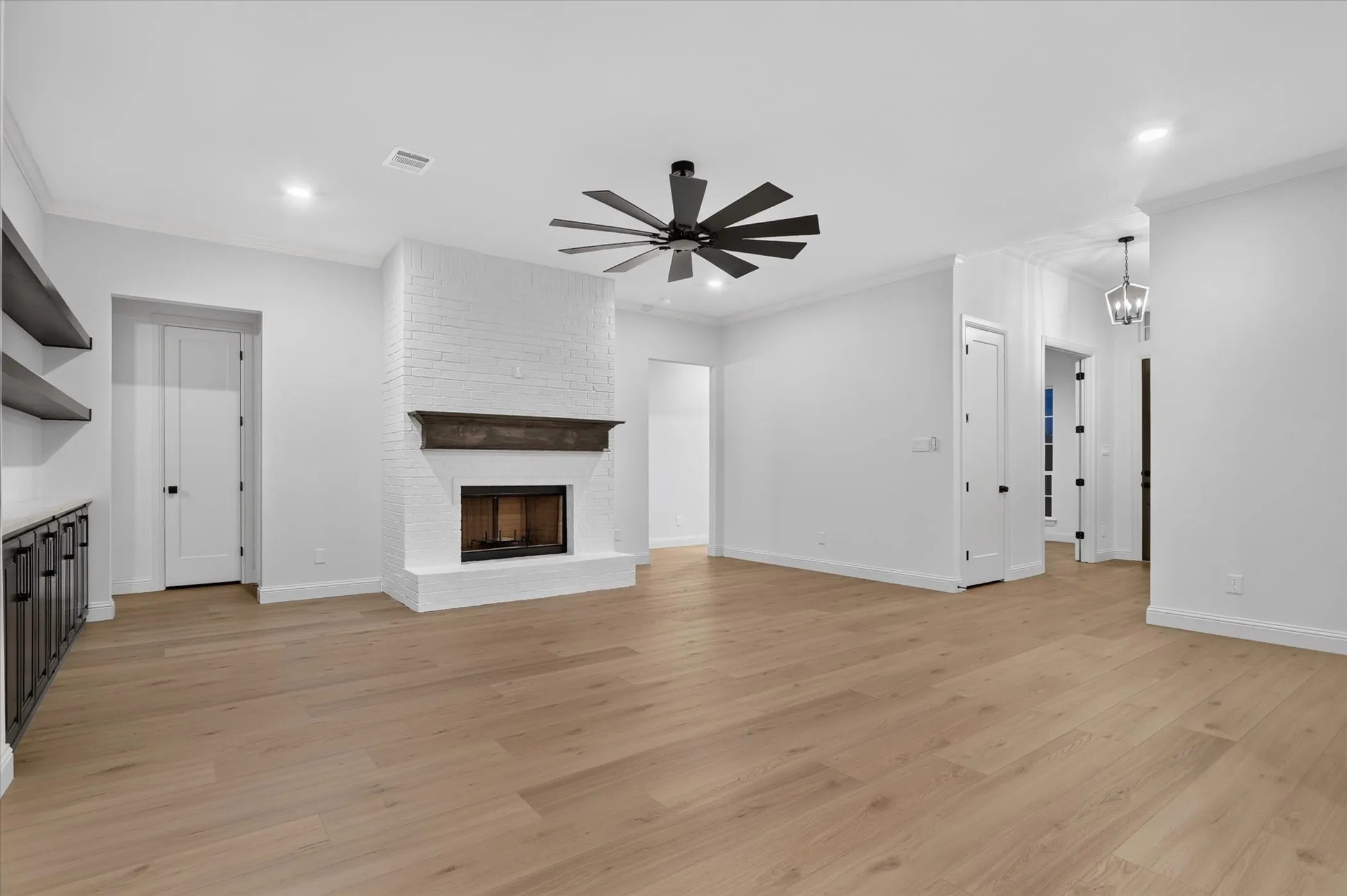 Light wood-finish flooring, white brick fireplace with dark wood mantel, built-in cabinetry with open shelving, contemporary ceiling fan, recessed lighting