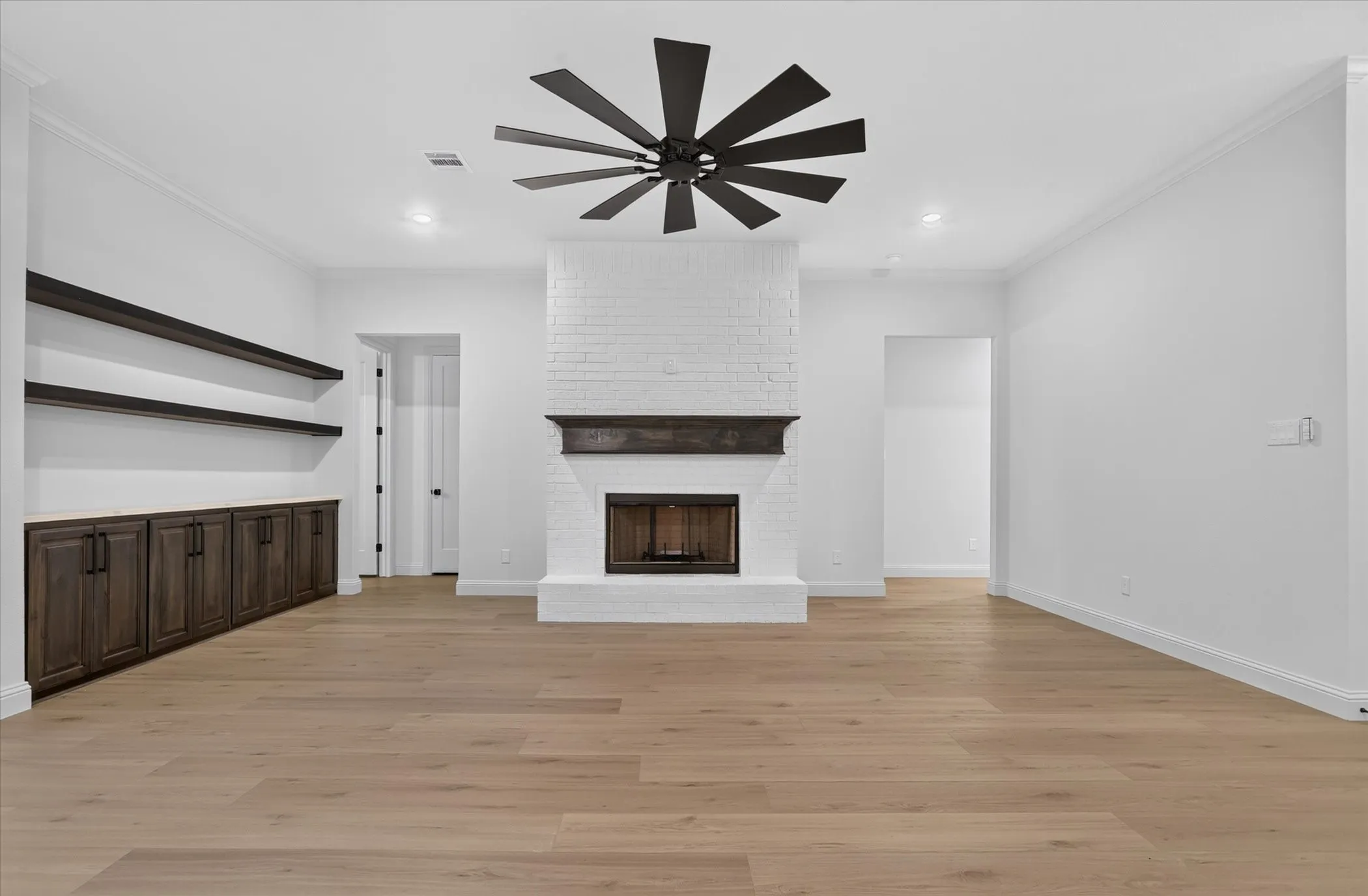 Wood-finish flooring, white painted brick fireplace with dark wood mantel, built-in cabinetry with dark wood finish, floating shelves, and a ceiling fan