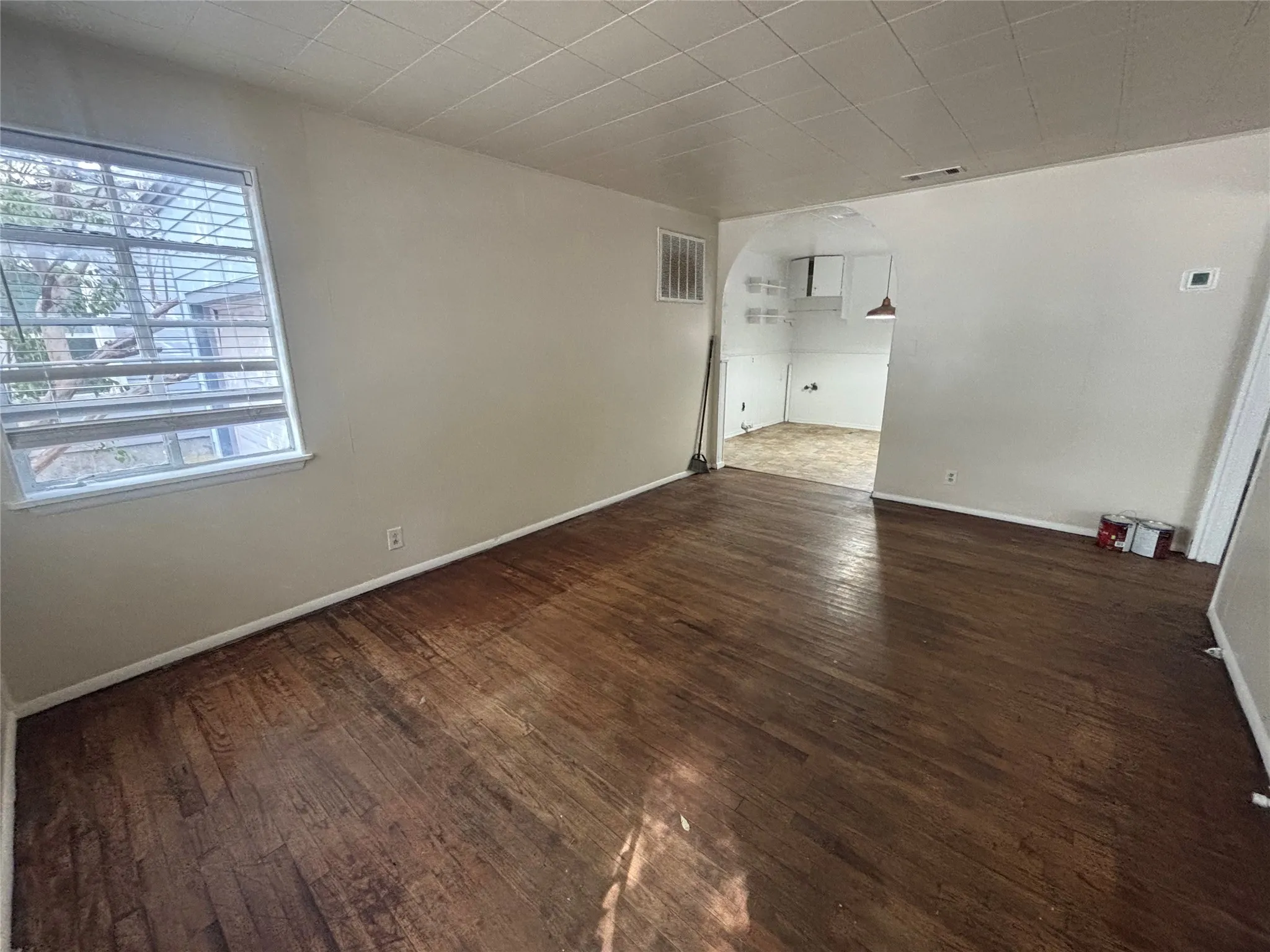Hardwood flooring, arched doorway, window with horizontal blinds, neutral wall paint, and ceiling with acoustic tiles