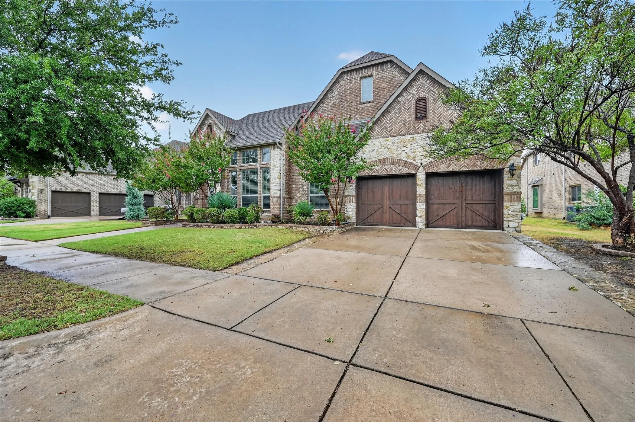 Brick and stone facade, arched garage doors, concrete driveway, and mature landscaping
