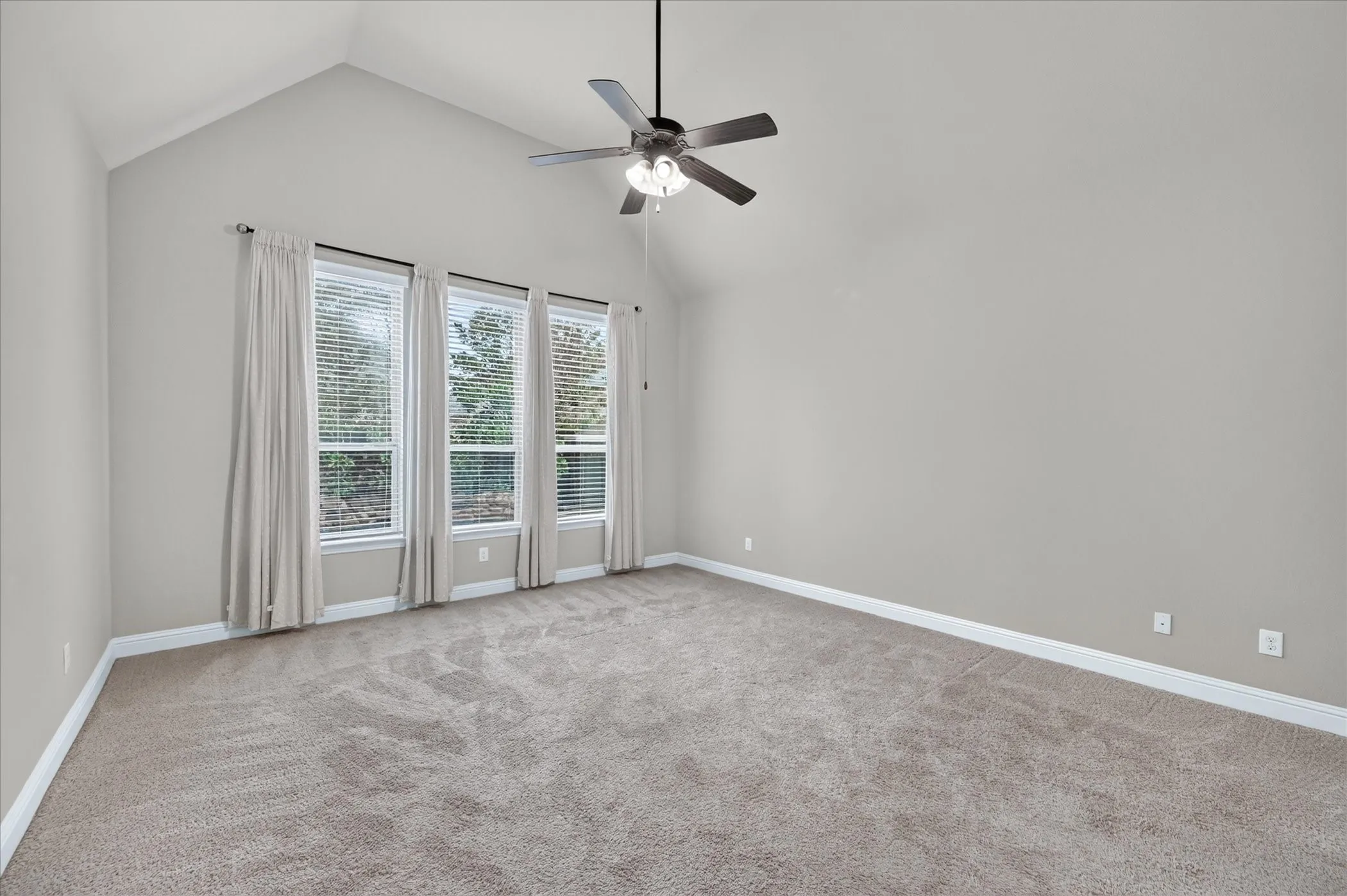 Vaulted ceiling with a ceiling fan, three large windows, and neutral carpeting