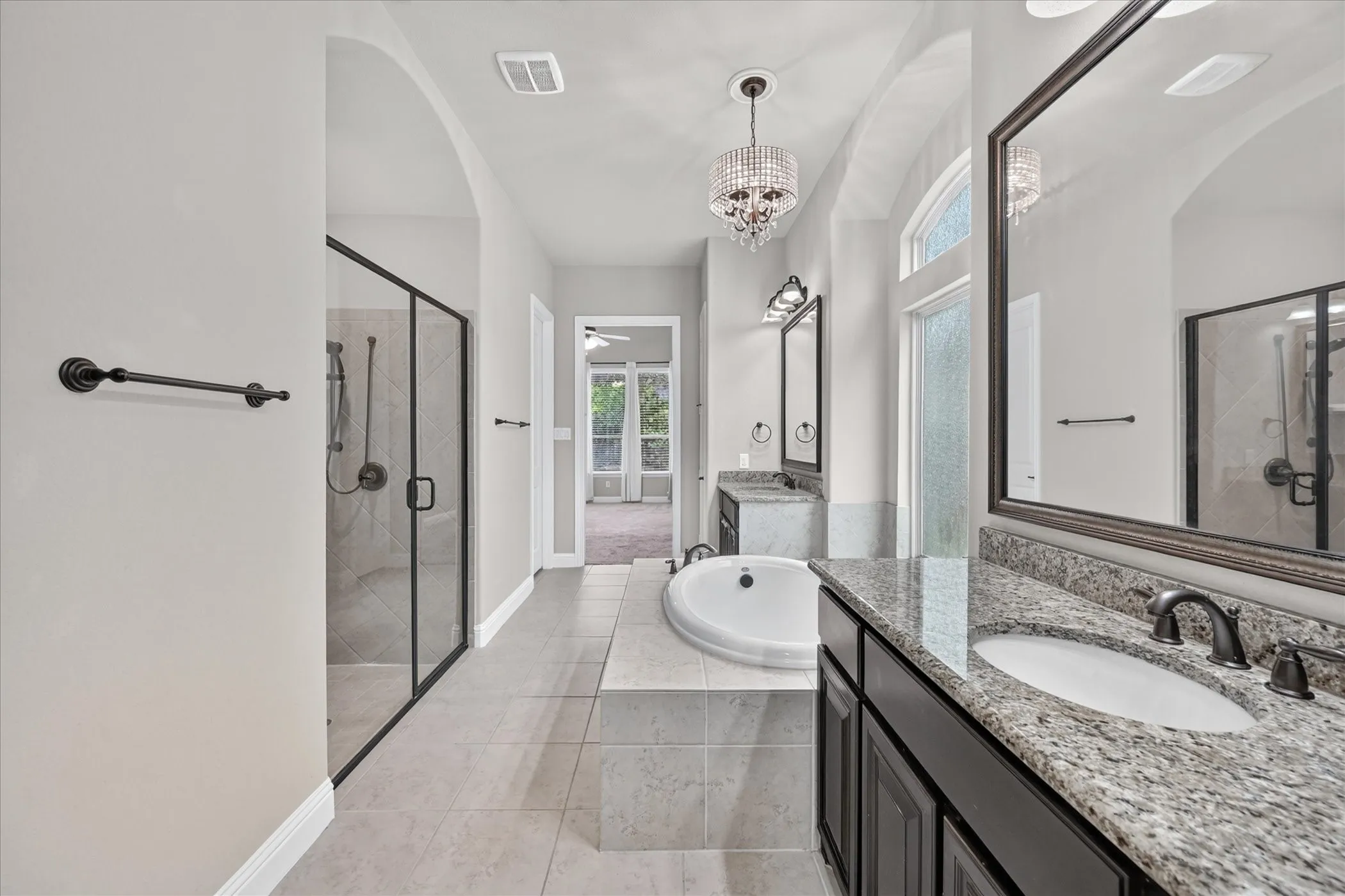 Dual vanity with granite countertops and undermount sinks, separate tiled shower with glass enclosure, soaking tub, and chandelier lighting
