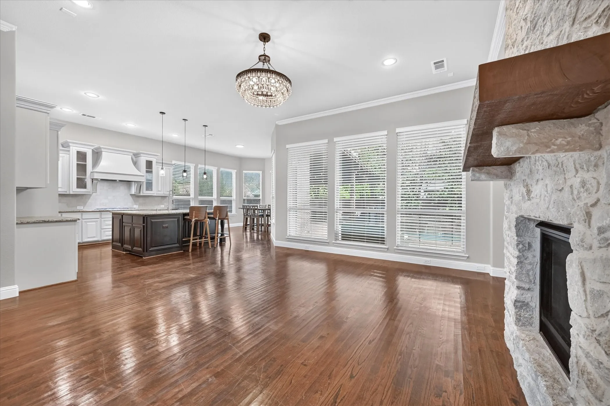 Hardwood flooring, stone-clad fireplace with wood mantle, multiple windows with blinds, built-in kitchen island, and crystal chandelier