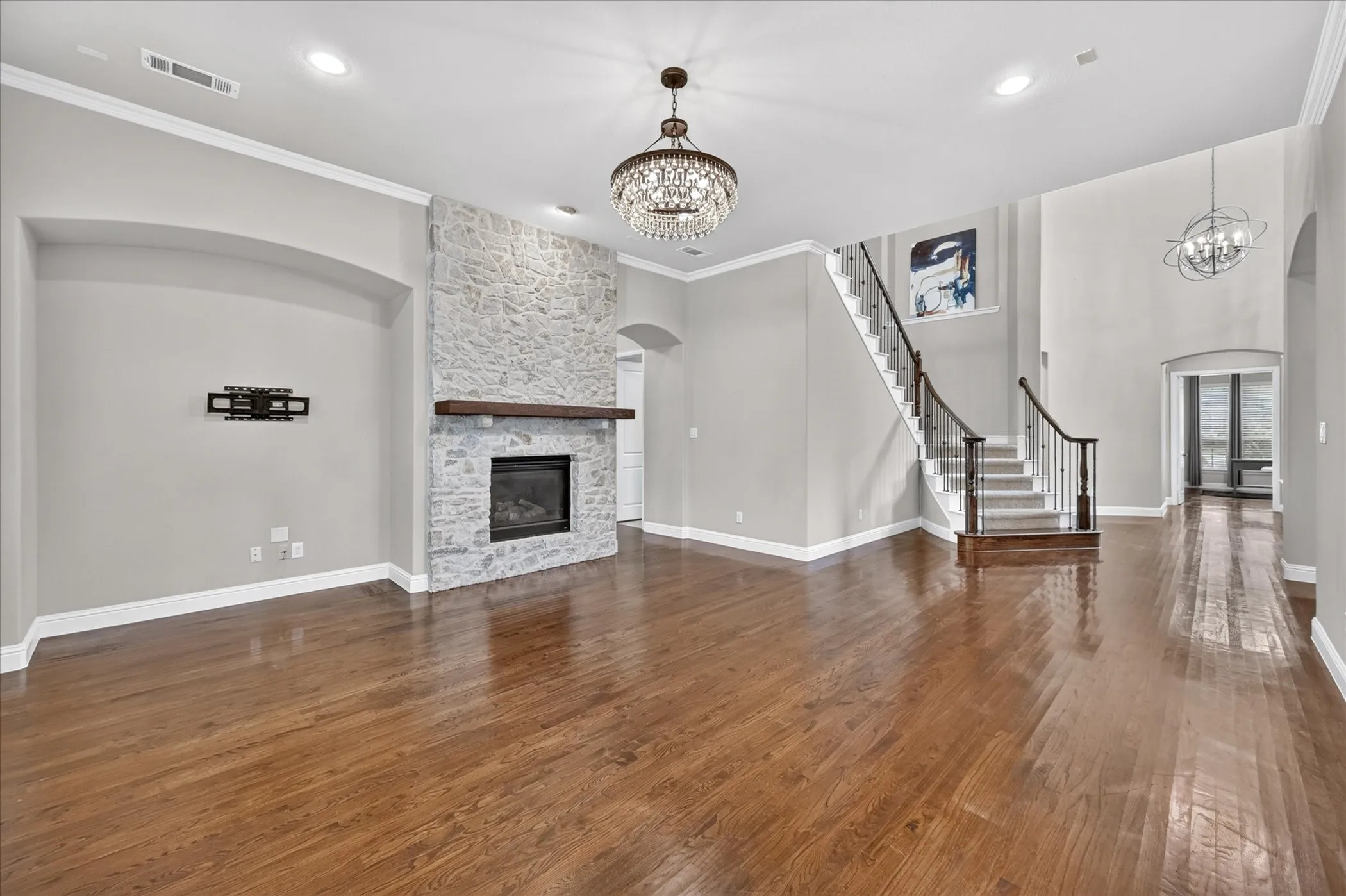 Hardwood flooring, stone-clad fireplace with wood mantel, crystal chandelier, recessed lighting, and a curved staircase with dark wood banister