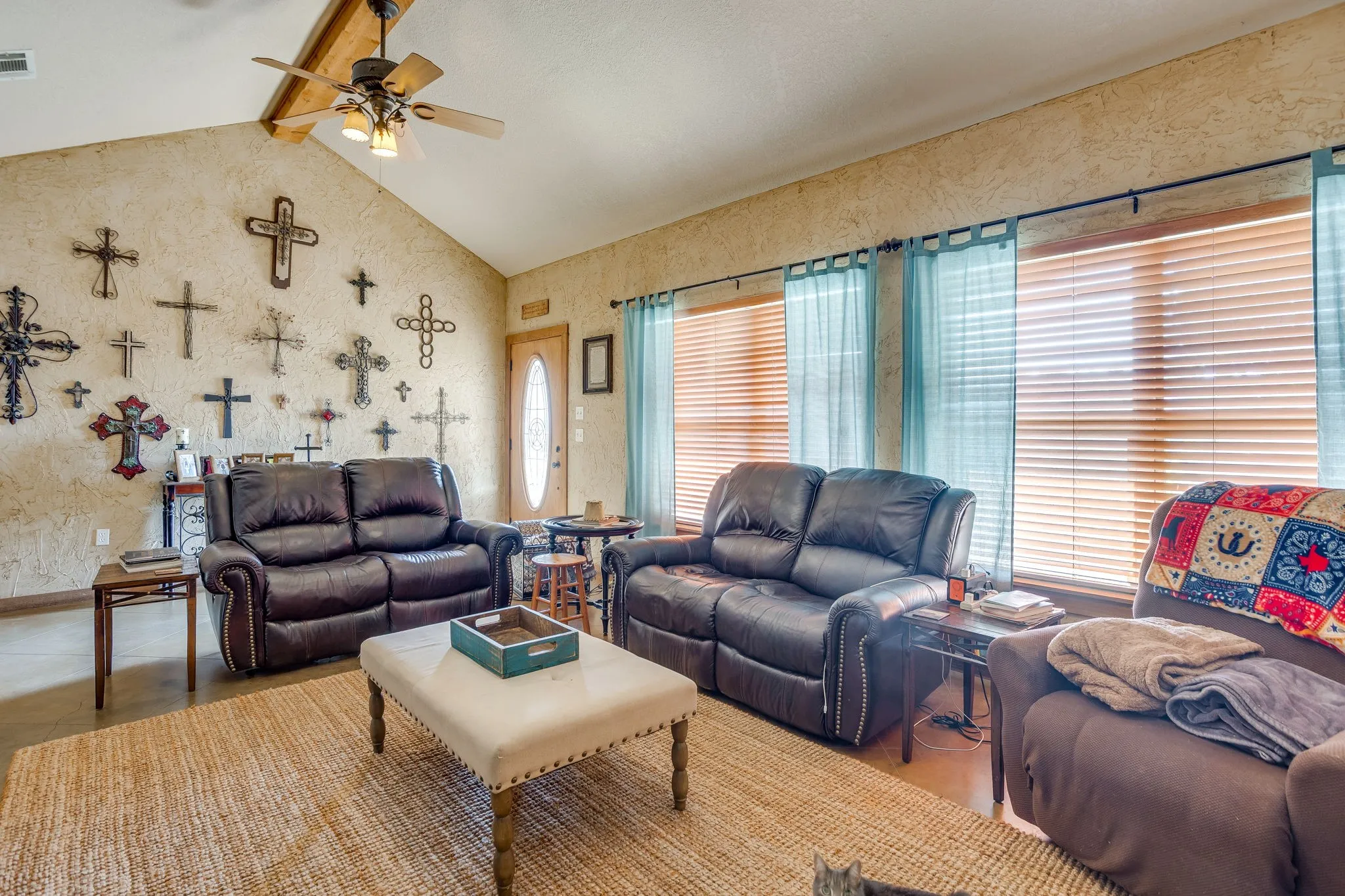 Vaulted ceiling with exposed wood beam, ceiling fan with light fixture, multiple windows with blinds, and a paneled entry door with decorative glass insert