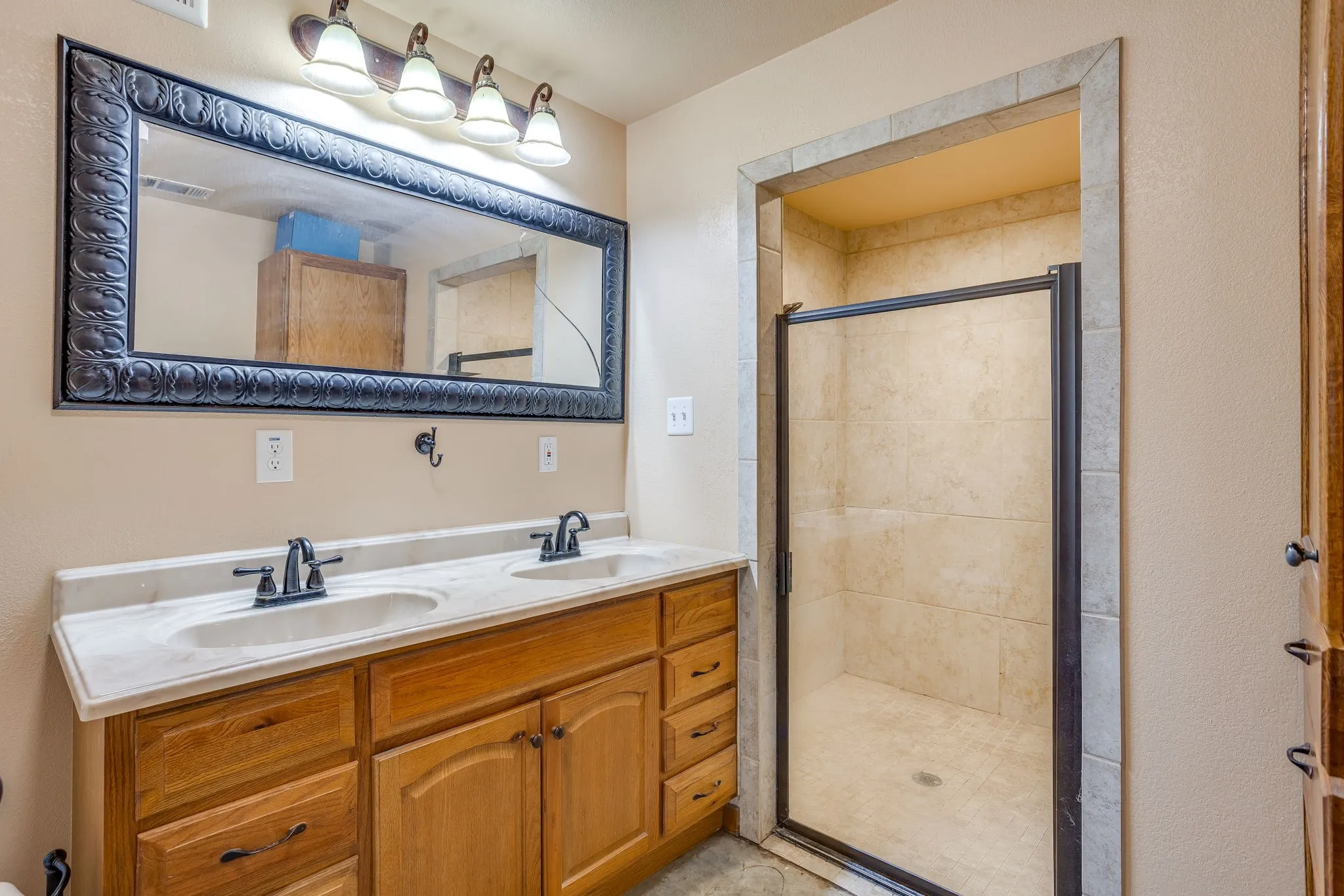 Dual vanity with undermount sinks and dark bronze fixtures, framed mirror, four-light vanity fixture, and a tiled walk-in shower with glass enclosure