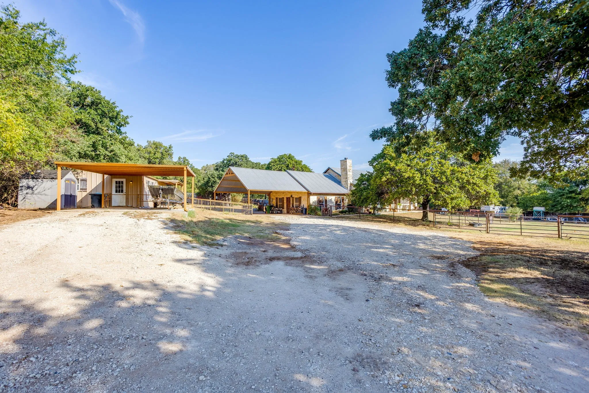 Gravel driveway leading to multiple structures, including a shed with an attached carport and a larger residence featuring a metal roof and stone chimney