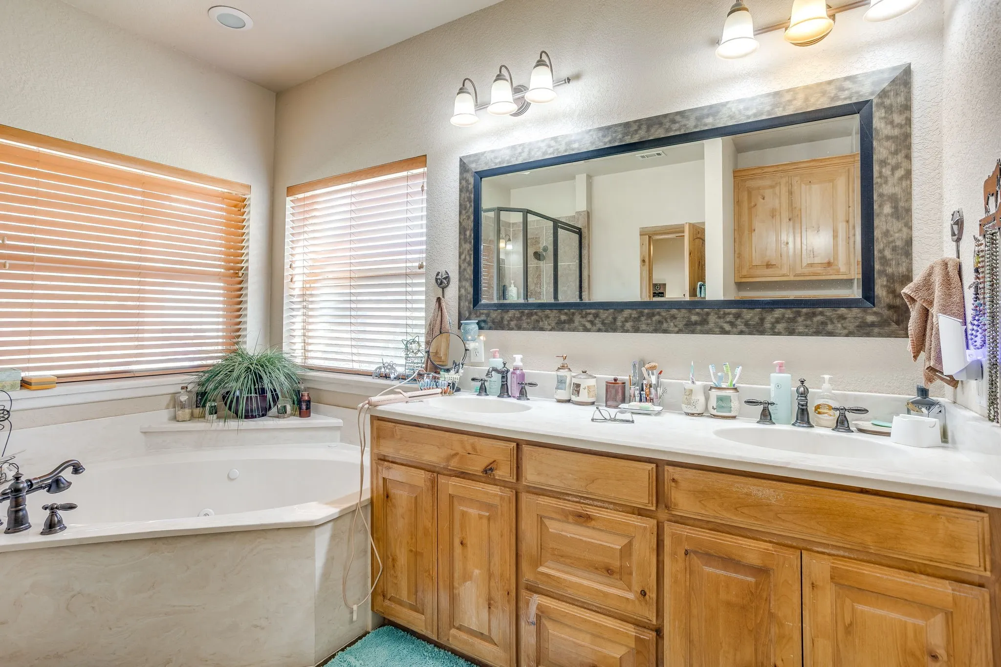 Bathroom featuring a corner soaking tub, dual windows with wood-finish blinds, a double vanity with integrated sinks, and a large framed mirror with overhead lighting
