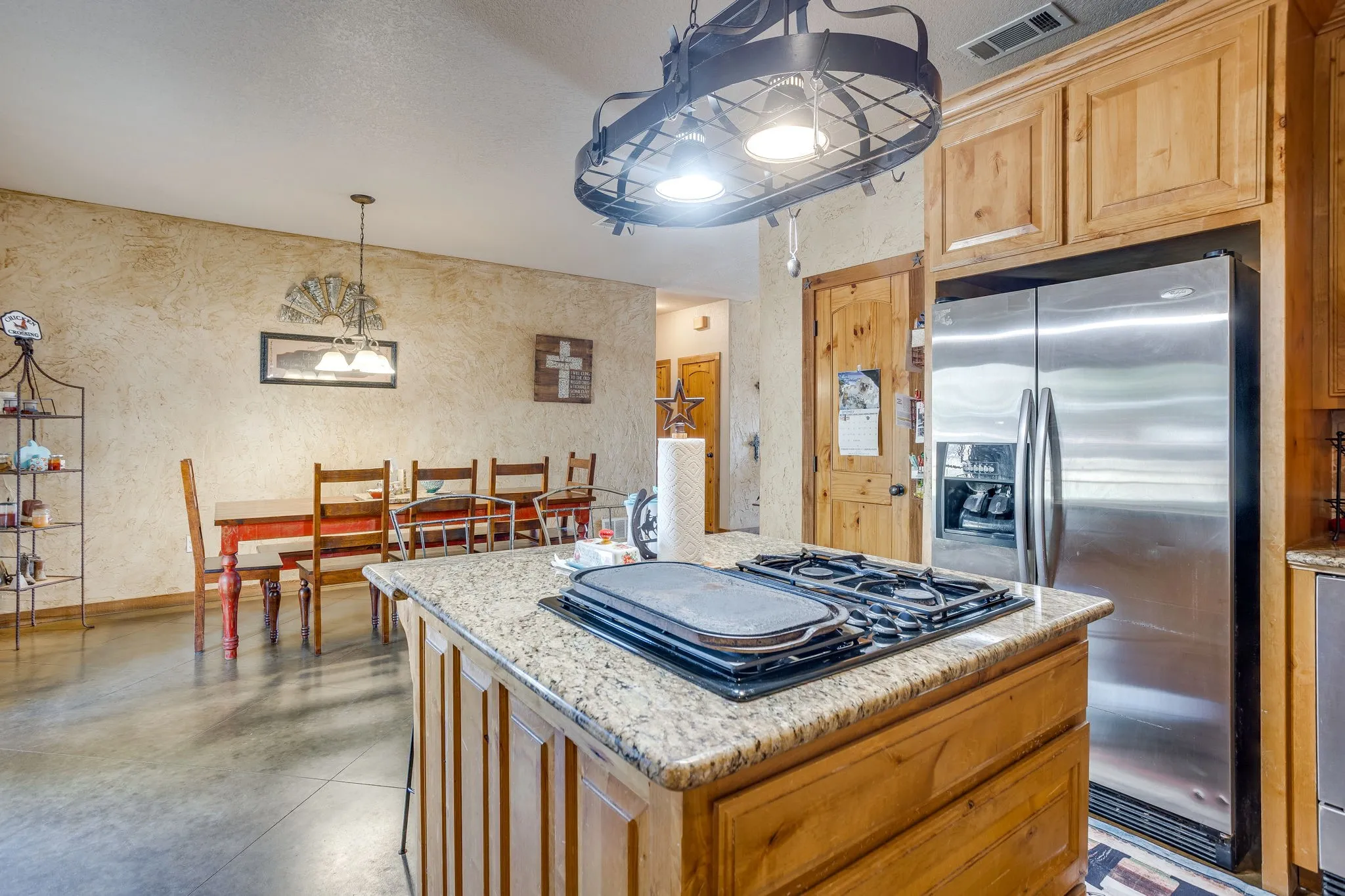 Kitchen island with integrated cooktop, granite countertop, stainless steel refrigerator, and wood​​‌​​​​‌​​‌‌​‌‌​​​‌‌​‌​‌​‌​​​‌​​ cabinetry