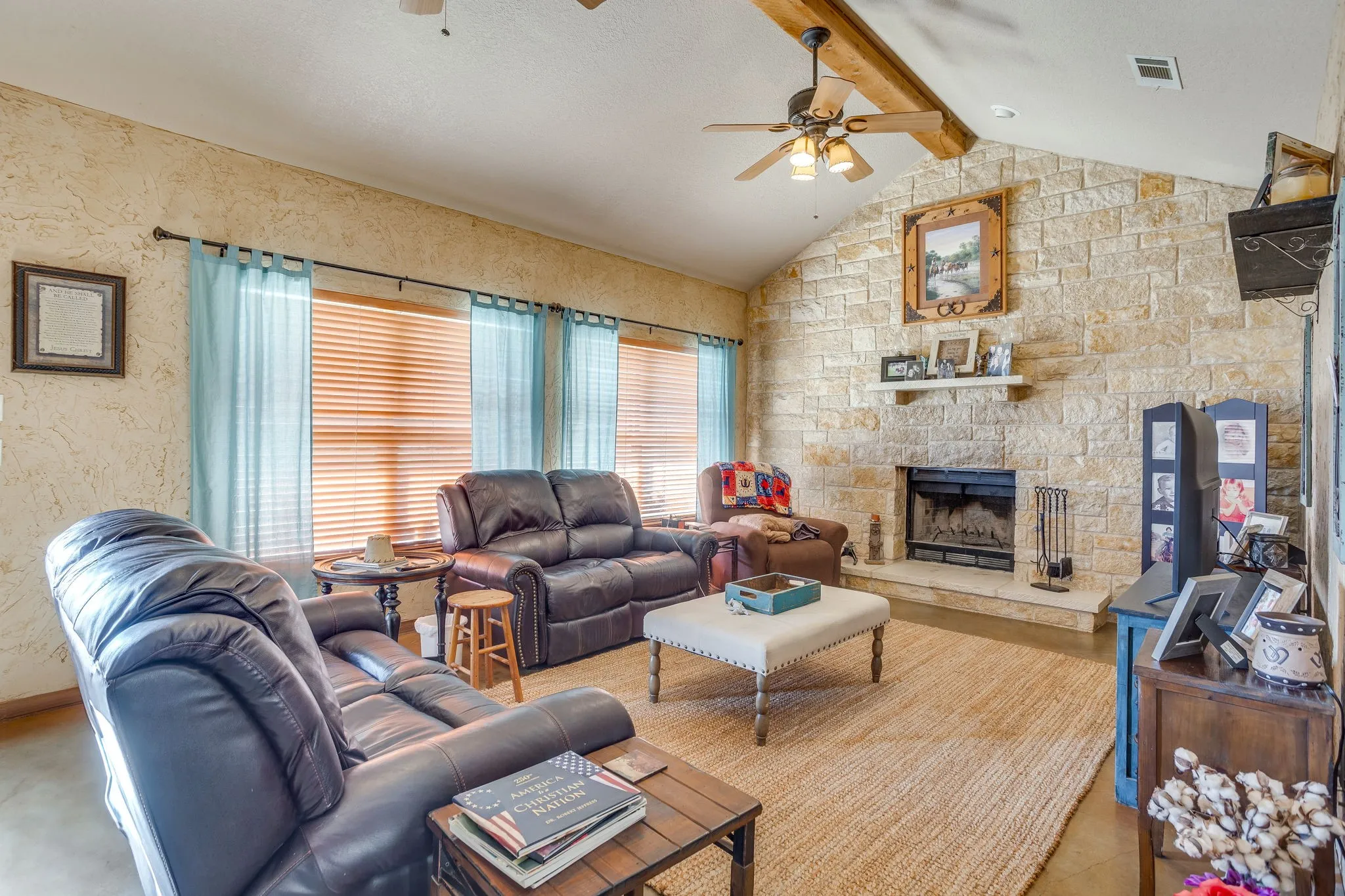 Vaulted ceiling with exposed wood beam, stone-clad fireplace, built-in fireplace insert, multiple windows with blinds, and ceiling fan with integrated lighting