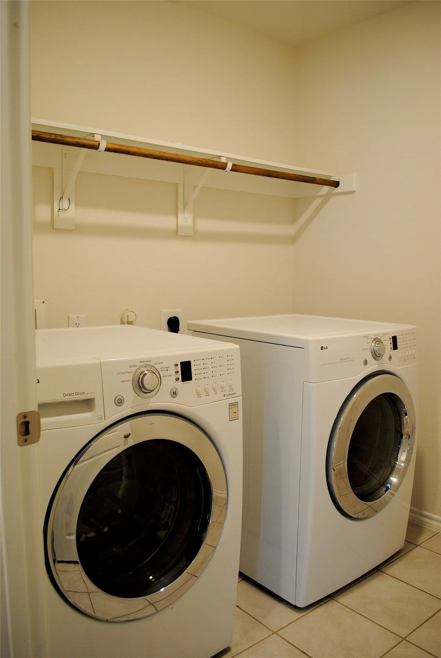 Dedicated laundry room featuring side-by-side front-load washers and dryers with stainless steel finishes, tile flooring, and built-in shelving with wooden accents for convenient storage and organization