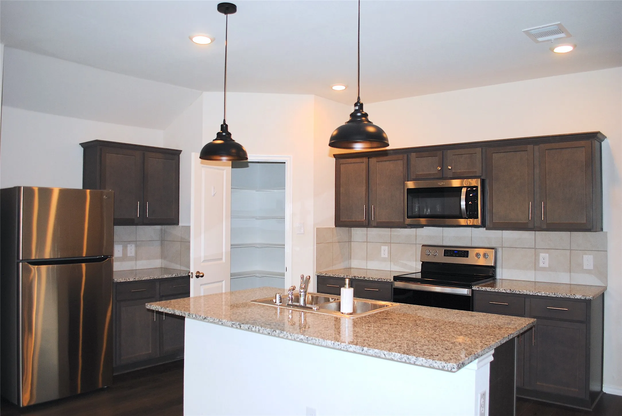 Kitchen featuring granite countertops, stainless steel appliances, dark wood-finish cabinetry, a tile backsplash, and a pantry with shelving