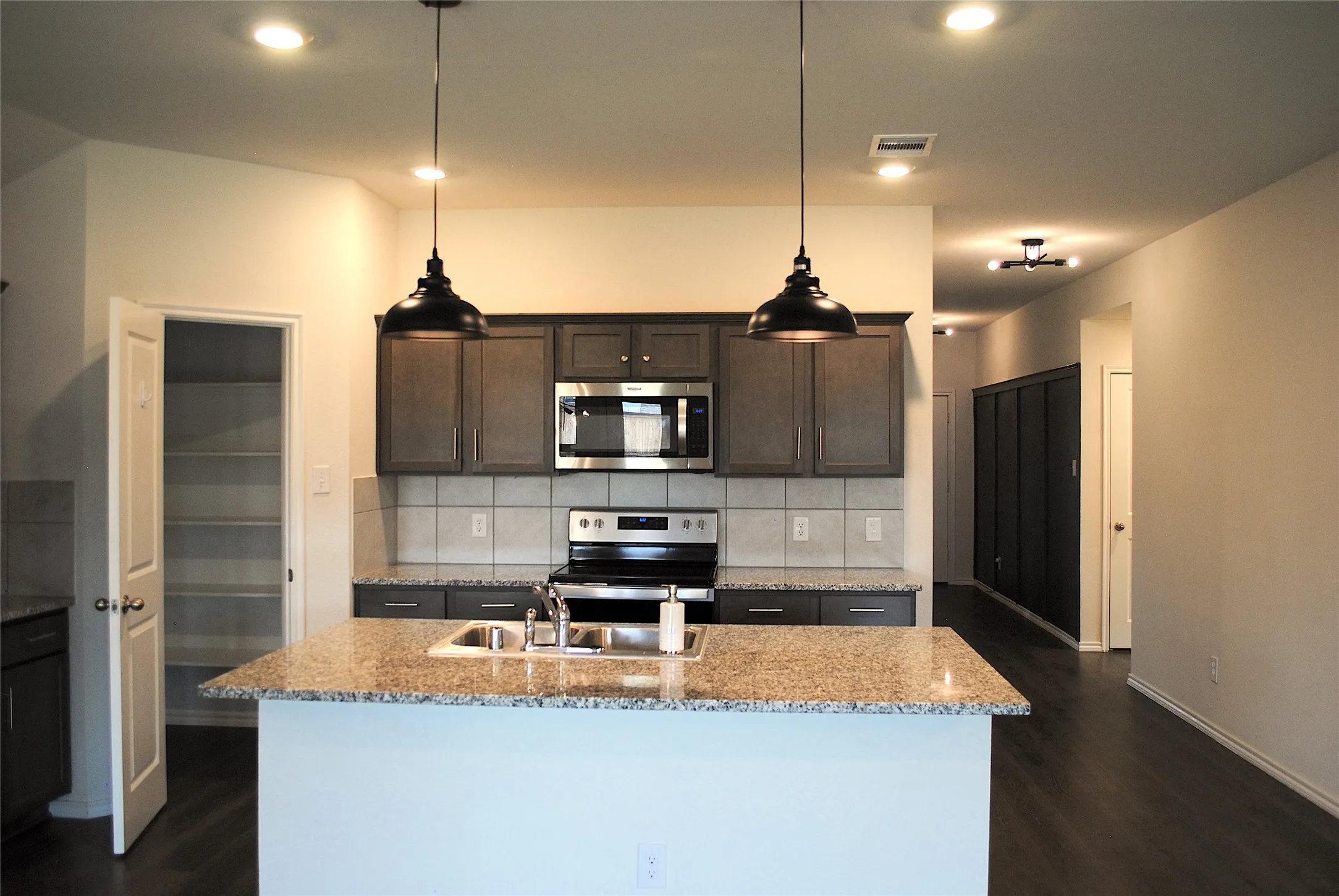 Kitchen featuring a center island with sink, granite countertops, stainless steel appliances, dark wood-finish cabinetry, and a tile backsplash