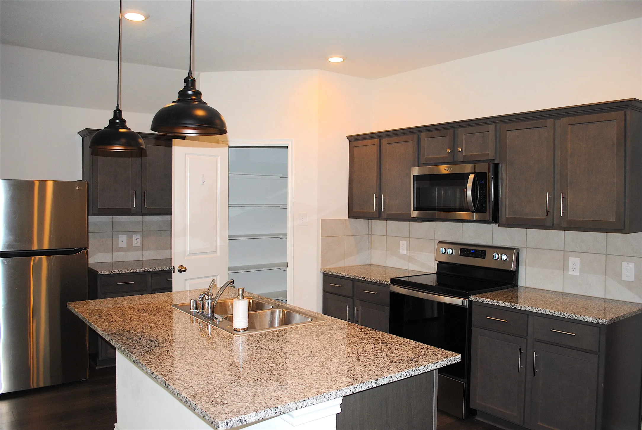 Kitchen featuring an island with a built-in sink, dark wood-finish cabinetry, light-toned countertops, tile backsplash, and stainless steel appliances