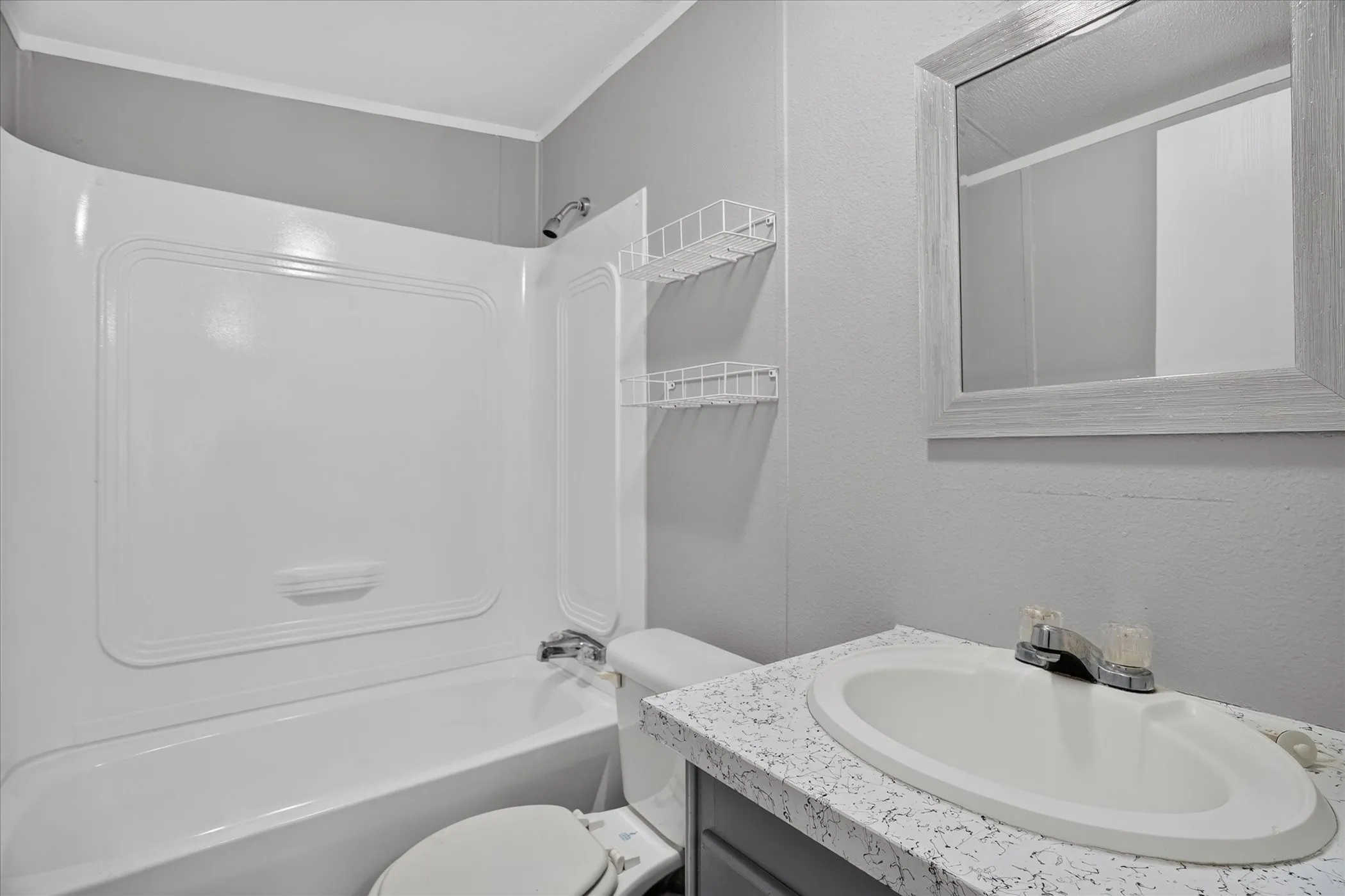 White integrated shower/tub unit with chrome fixtures, inset oval sink with chrome faucet, speckled countertop, and framed vanity​​‌​​​​‌​​‌‌​‌‌​​​‌‌​‌​‌​‌​​​‌​​ mirror