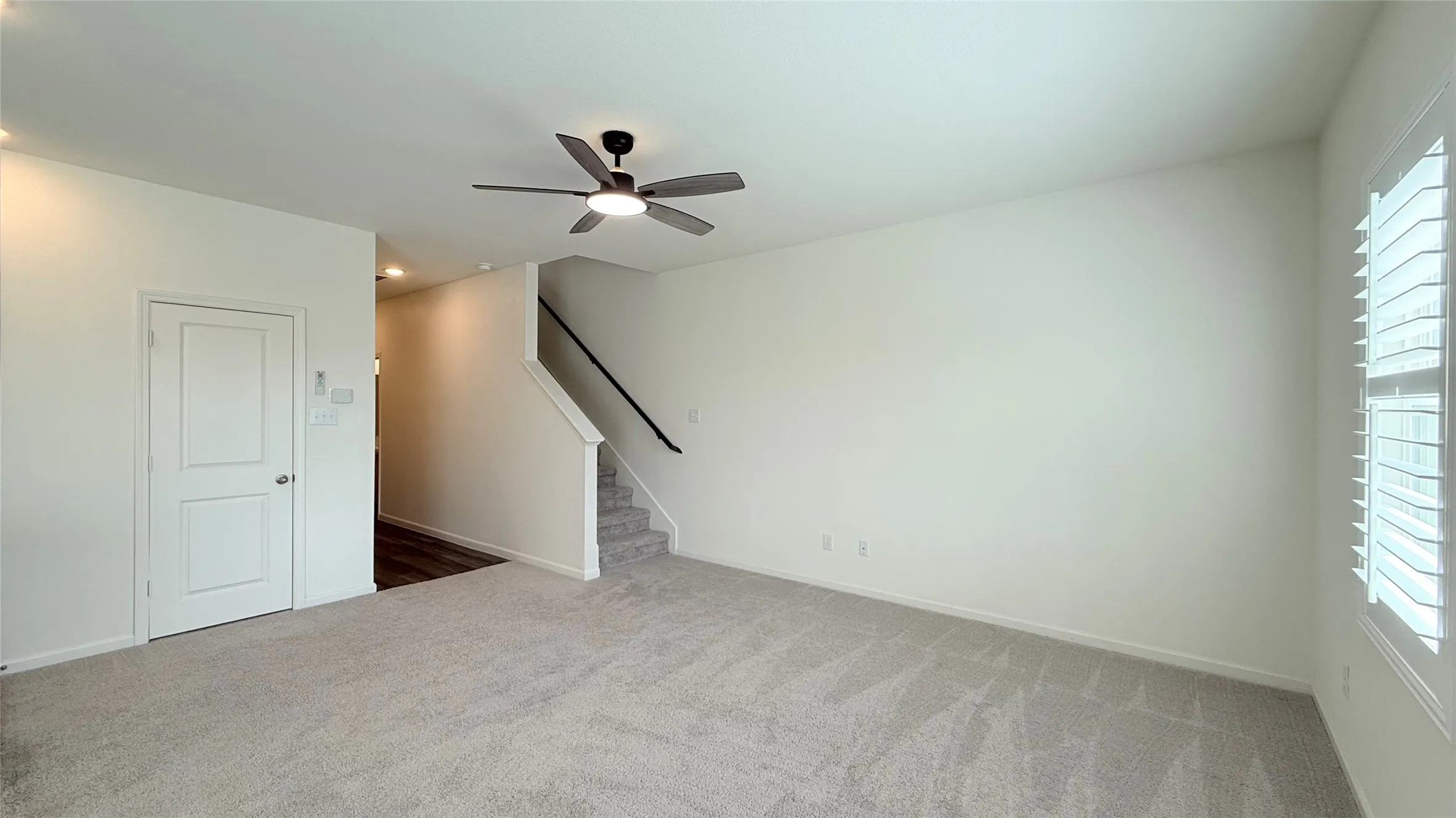 Open-concept living space featuring light-toned carpeting, a ceiling fan with integrated lighting, and a window with plantation shutters