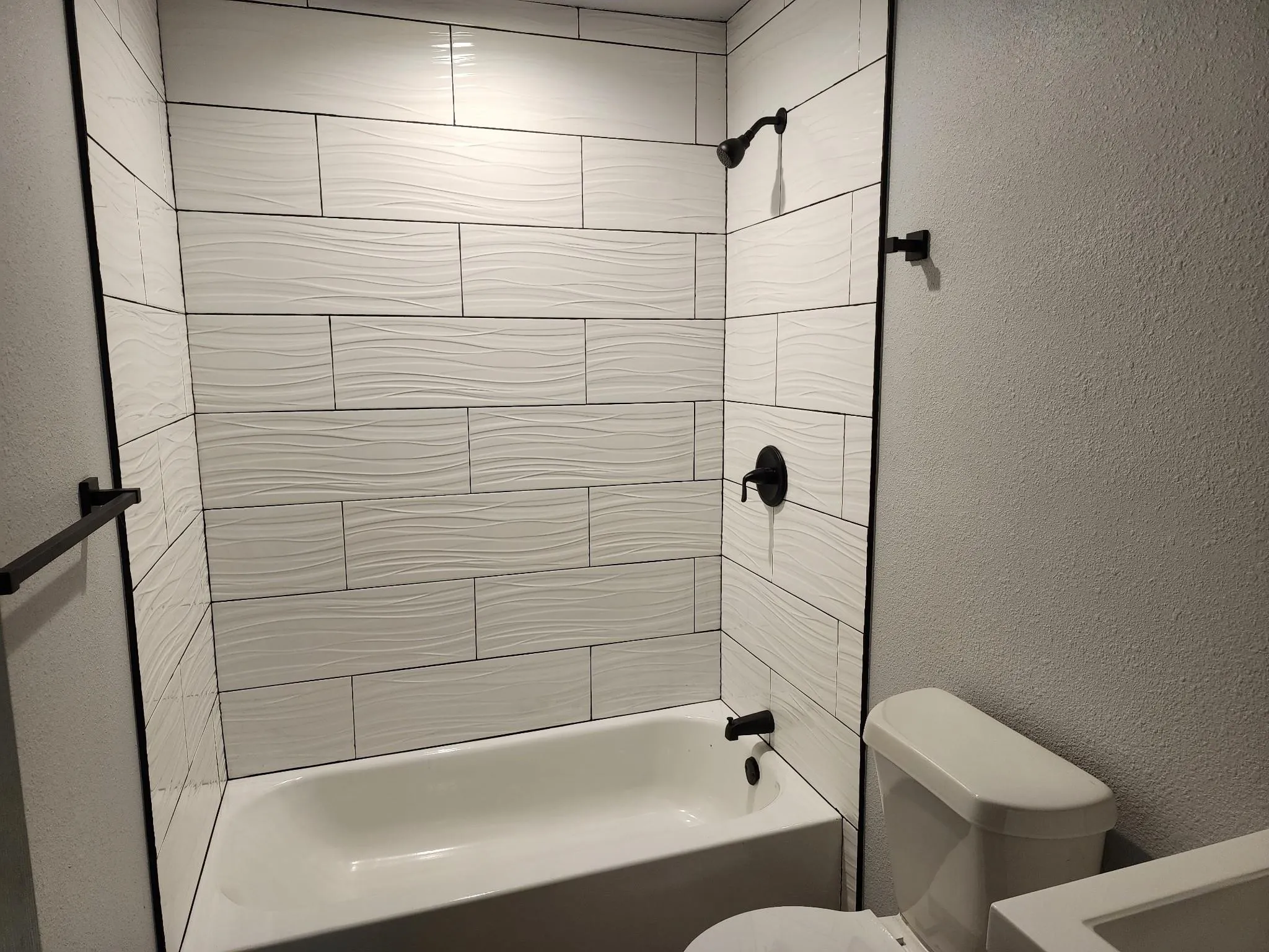 Tub-shower combination with textured white tile surround, matte black fixtures, and a white porcelain toilet